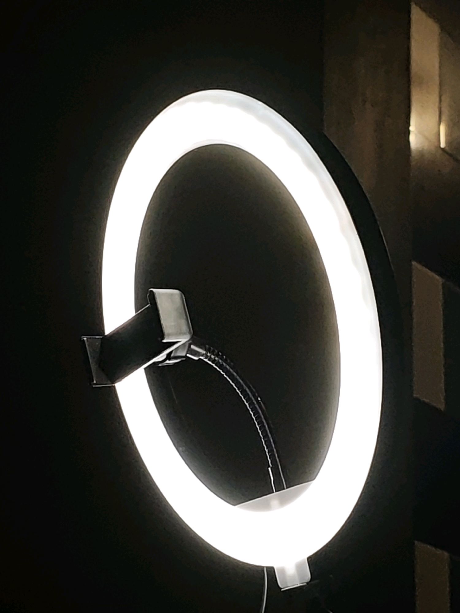 A ring light is illuminated against a dark background, with a dark building visible to the right. The ring light has a metallic bracket attached to its side, which is connected to a flexible gooseneck arm. The bracket is silver and appears to be made of metal. The gooseneck arm is black and is slightly bent. The ring light is circular and emits a bright white light. The building to the right is dark and appears to have some vertical lines or indentations. The scene is a close-up shot of the ring light, with the background blurred. There are no people or animals visible in the image. The setting is likely indoors, or outdoors at night, in Ngasem, Indonesia.