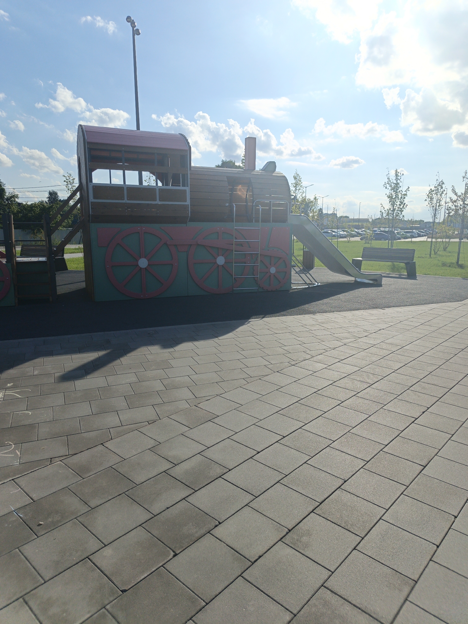 The image shows a modern playground structure resembling a train, located in a paved public area. The train structure is made of wood and metal, featuring a red roof, brown sides with large green wheels outlined in red, and a red smokestack. A silver ladder is attached to the side, leading up to a sliding board which is a bright green. In the background, there is a parking lot filled with cars, trees, streetlights, and a cloudy blue sky with bright sunlight. A park bench is visible near the sliding board. The foreground is dominated by a grey tiled walkway. The scene is devoid of people. The image was taken on a sunny day. Based on the context provided, the location is identified as Pavlovsky Posad, Russia.