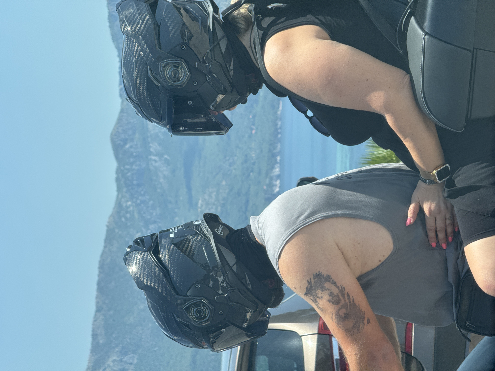 Two individuals wearing black motorcycle helmets are seated on a vehicle, viewed from the rear and right side. The person on the left has a tattoo on their left arm and is wearing a gray tank top. The person on the right is wearing a black tank top. The right person's left hand is resting on the left person's torso. The background features a body of water and mountains, consistent with the coastline near Budva, Montenegro. The lighting suggests a sunny day.