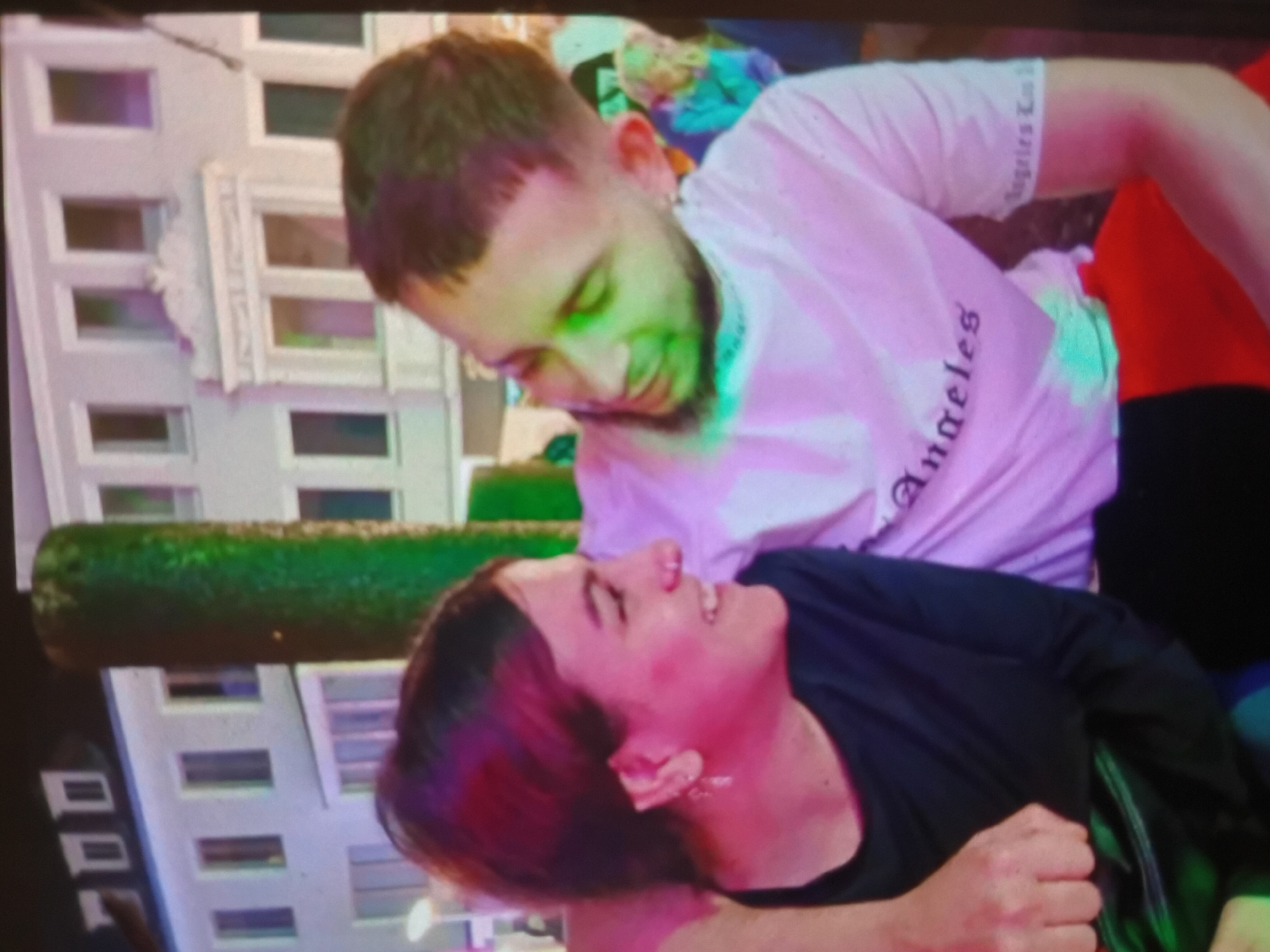 The image shows a close-up of a young couple embracing and looking at each other. The woman, on the left, has brown hair and is wearing a dark top. She is smiling and has rosy cheeks. The man, on the right, has short dark hair and is wearing a white t-shirt with "Angeles" printed on it. He is also smiling and appears to be looking at the woman. They are in an outdoor setting with a building in the background. The building appears to be a multi-story structure with several windows. There is also a tall, green, cylindrical object in the background, possibly a decorative pillar or a tree. The overall lighting suggests it is evening or night, with artificial lights illuminating the scene. There is no indication of Hrishteni, Bulgaria in the image.