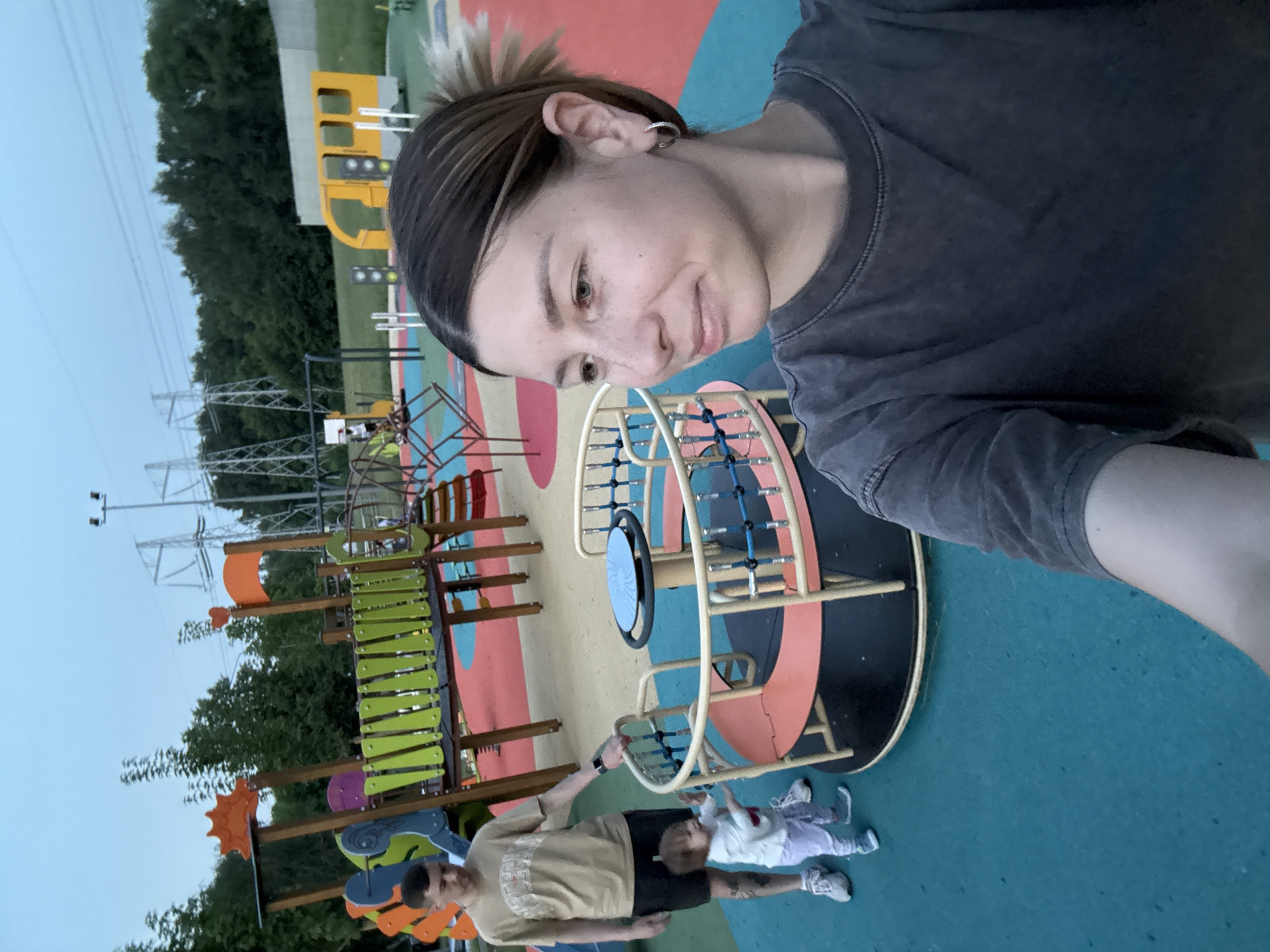 This is a selfie taken at a modern, colorful playground in Moscow, Russia. The playground features a yellow train-shaped structure, a bright orange and green climbing frame, and a circular merry-go-round with a blue disc. In the foreground, a woman with light brown hair pulled back is looking to her left, wearing a dark grey t-shirt. A man in a tan t-shirt and black shorts stands near the merry-go-round, with his hand on its railing. A small child in a white top and light purple pants is also near the merry-go-round, appearing to interact with it. The ground of the playground is a safety surface in various colors, including teal and red. In the background, there are trees and large electrical pylons, suggesting an urban or suburban setting. The sky is overcast and the lighting indicates it is either early morning or late evening.