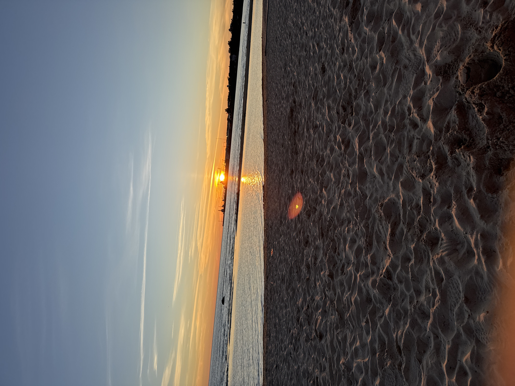 This photograph captures a sunset over the Baltic Sea at a beach in Tallinn, Estonia. The foreground is dominated by the textured sand of the beach, with gentle ripples and shadows cast by the low-angled sunlight. The water of the sea forms the middle ground, appearing calm with reflections of the sky and sunset. In the distance, along the horizon, a silhouette of structures is visible, possibly industrial cranes and buildings, suggesting a port or industrial area. The sky exhibits a gradient of colors, transitioning from a pale blue at the top to warm oranges and yellows closer to the horizon where the sun is setting. Thin, wispy clouds streak across the sky. A bright lens flare, appearing as a warm orange circle, is present in the lower right quadrant of the image. No individuals or specific actions are discernible in the scene.