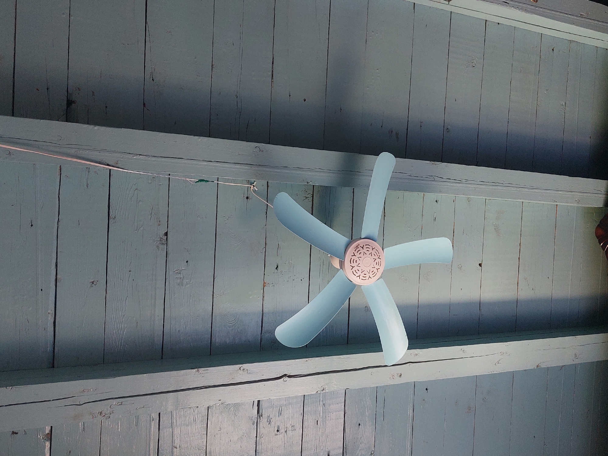 A pale blue ceiling fan with five blades hangs suspended by a white cord from a wooden plank ceiling. The ceiling is composed of horizontal wooden planks painted a light blue color, with visible grain and imperfections. Two thick wooden beams, also painted light blue, run diagonally across the image, dividing the ceiling into sections. One beam is on the left and the other is on the right. The fan itself has a pinkish-beige central housing with a circular, decorative pattern. The overall lighting suggests an indoor setting, possibly with natural light coming from an unseen source, casting shadows from the beams onto the ceiling. The scene is static and depicts an object and its surroundings.