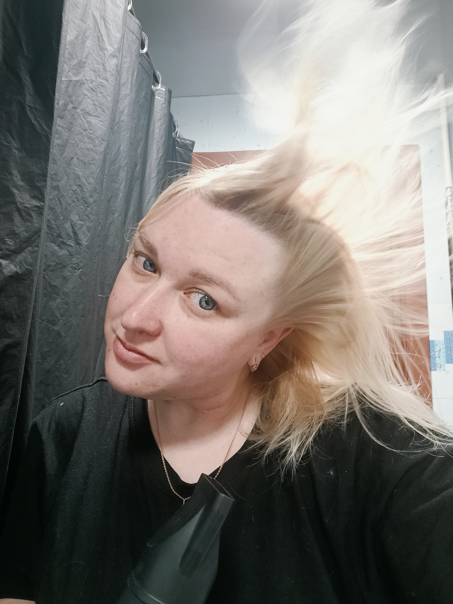 A woman with fair skin, blue eyes, and blonde hair is holding a hair dryer in front of her in a bathroom. Her hair is being blown upward by the dryer. She is wearing a black t-shirt and a delicate gold necklace. A black shower curtain hangs in the background to the left, and a wall with partially visible tiles is to the right. The photograph appears to have been taken at a close distance, focusing on the woman's face and hair. The location is Chaikovsky, Russia.