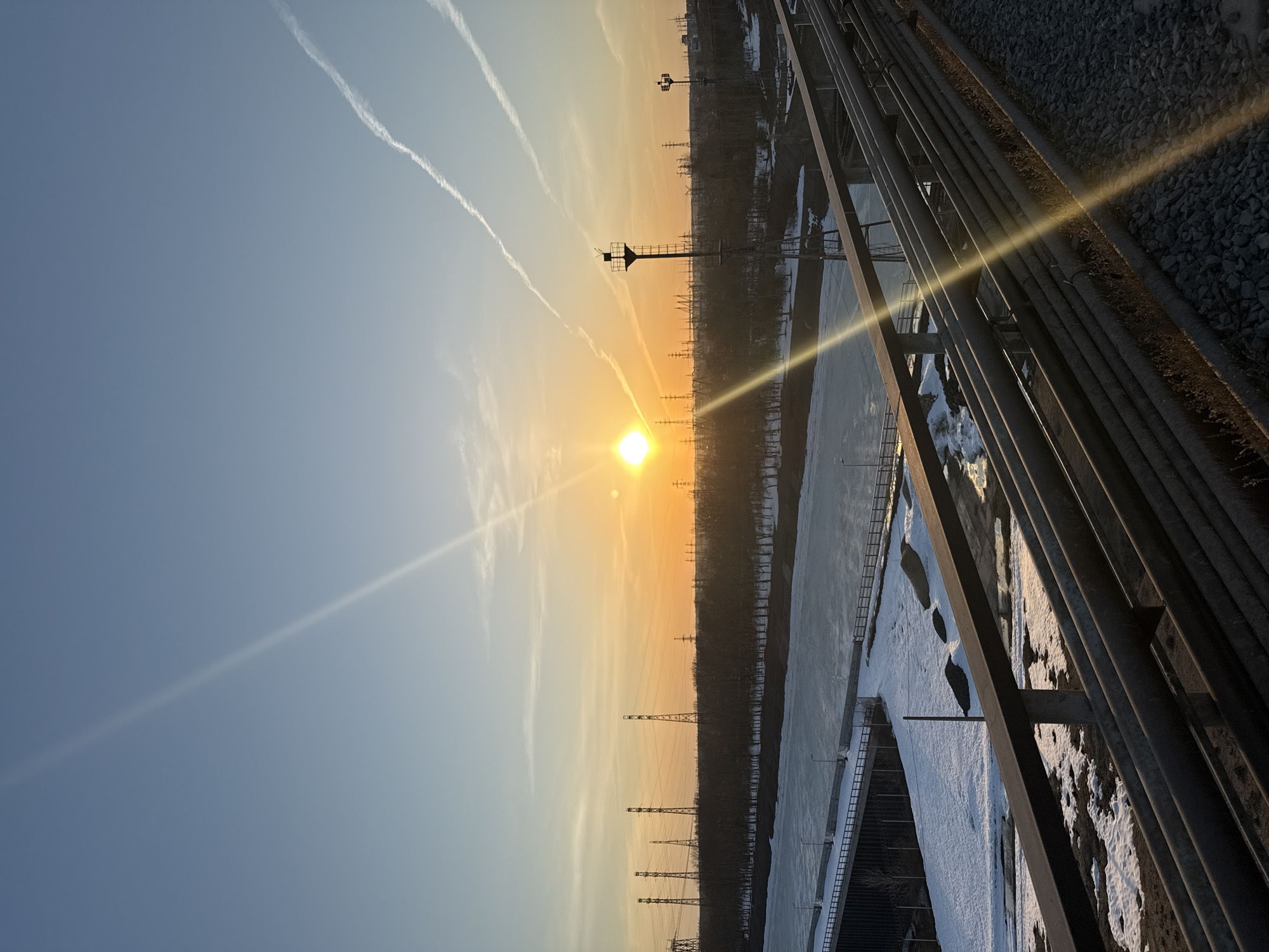 The photograph depicts a sunset scene near an industrial area in Chaikovsky, Russia. In the foreground, metal pipes and structural elements are visible, likely part of an industrial facility or bridge. Adjacent to these structures, there is a partially frozen body of water or wetland with snow-covered areas. A line of electrical transmission towers stretches into the distance along the horizon. Two contrails are visible in the sky above the sunset. The sky transitions from a light blue in the upper part of the image to a warmer, orange hue closer to the setting sun.
