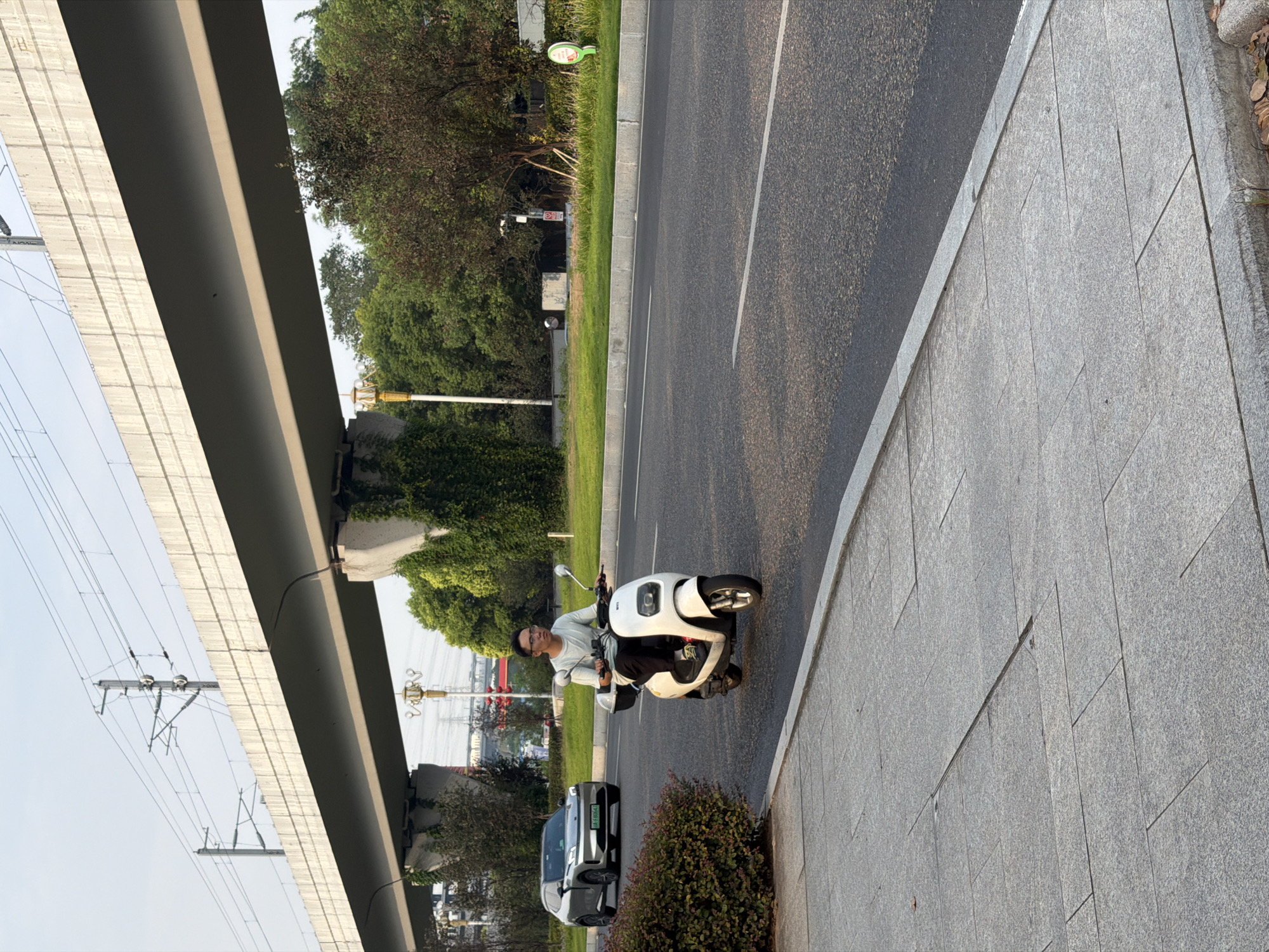 A man wearing glasses and a light-colored t-shirt rides a white electric scooter on a paved road in Jiangjin, China. He is on the right side of the road, approaching from the viewer's left. To his left, a silver SUV is driving in the same direction. The road is lined with a grey concrete sidewalk on the left and a grassy area with trees on the right. An elevated concrete structure, likely an overpass for a railway or highway, spans across the top of the image, with power lines visible above it. The sky is clear and bright.