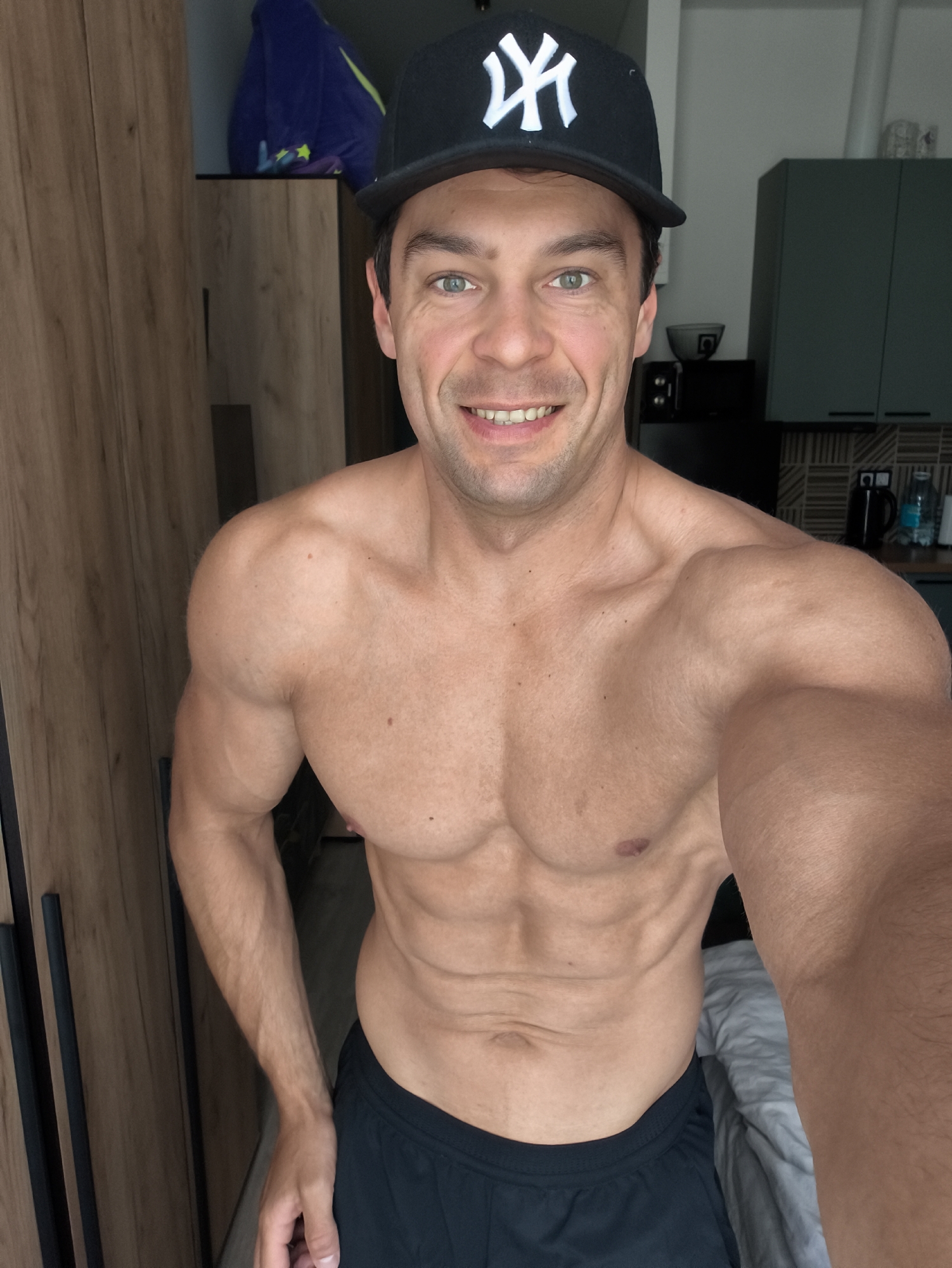 A man is posing for a selfie in a room, possibly an apartment. He is shirtless, wearing a black baseball cap with a white "NY" logo. He has a muscular physique with visible abs. He has green eyes and is smiling. The background shows a closet with wooden doors and a glimpse of a kitchen area with cabinets and appliances. There is also a bed with white and gray bedding visible. The lighting appears to be natural, coming from the right side of the frame. The overall impression is that of a casual, personal photo. The location is not specified in the image.