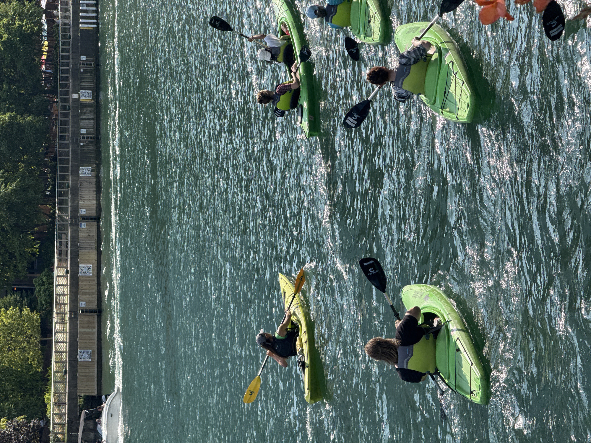 A group of six individuals are kayaking on a body of water. They are all in separate kayaks and appear to be paddling in the same direction. The kayaks are green, except for one that is yellow. The individuals are wearing life vests. In the background, there is a walkway with trees behind it. A boat is also visible on the water in the distance. The water is choppy and reflects the sunlight. The scene appears to be taking place on the Chicago River.