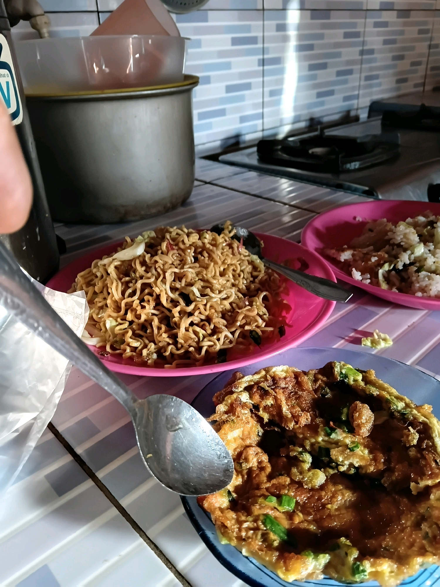 The image shows a close-up view of a table set with food, likely in a kitchen or dining area. In the foreground, a spoon rests on a blue plate containing a portion of what appears to be an omelet with green onions. Behind this, a pink plate holds a pile of instant noodles with some cabbage. To the right of that, another pink plate contains a portion of rice with vegetables. In the background, a metal pot with a plastic lid sits on a tiled surface. The overall impression is a casual meal setting, possibly in Indonesia given the mention of Margo Rukun. The lighting suggests indoor ambient light, with some glare from overhead light sources. The tiled backsplash behind the stove is visible, with a reflective sheen.