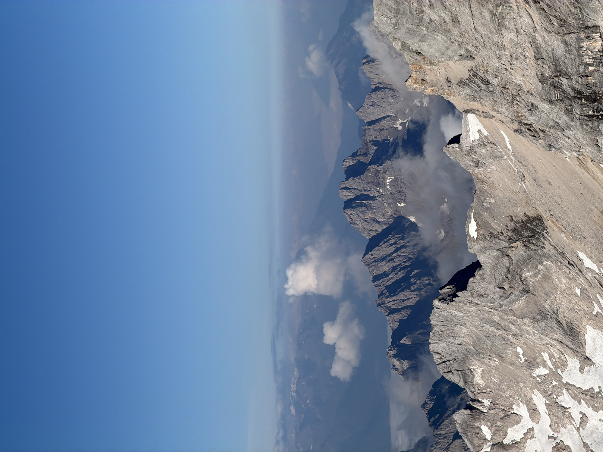 This is a high-angle, medium shot of a mountain range in Grainau, Germany, under a clear blue sky. The foreground is dominated by rugged, rocky terrain with patches of snow. Jagged peaks rise in the midground, partially obscured by wisps of clouds and mist. In the background, a hazy, distant mountain landscape stretches out, also veiled by atmospheric haze and clouds. The scene appears to be captured during daylight, with sunlight illuminating the mountainous terrain and casting shadows. No people or man-made objects are visible in the image.