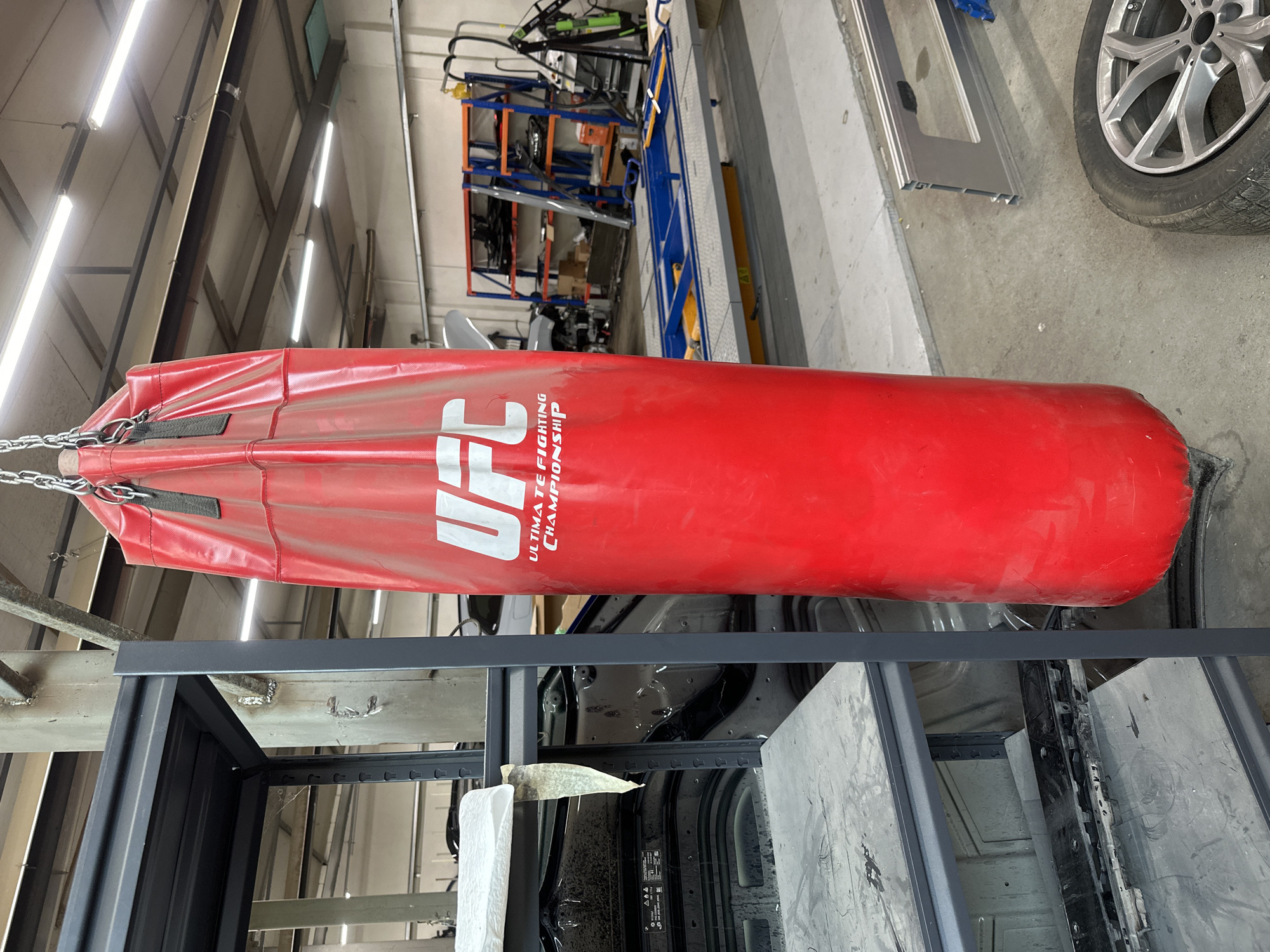 The image captures a large, red punching bag prominently displayed in what appears to be an industrial or workshop setting. The punching bag is adorned with the white "UFC" logo, accompanied by the text "ULTIMATE FIGHTING CHAMPIONSHIP" below it, and is suspended by metal chains.
The background features a high ceiling with exposed metal beams and numerous fluorescent light fixtures, illuminating the space. To the right, various industrial elements are visible, including blue and orange shelving units holding tools and equipment, a car wheel with a tire, and a silver metal frame resting on a concrete floor. The lower portion of the image shows black metal frames and what seems to be components of a vehicle's interior or chassis, suggesting an automotive repair shop, factory, or similar workspace.
No people are present, and no particular activity or event is taking place; the scene is static. The bright artificial lighting indicates an indoor setting, likely during daylight hours. The overall environment conveys a functional, utilitarian atmosphere without any discernible emotions.