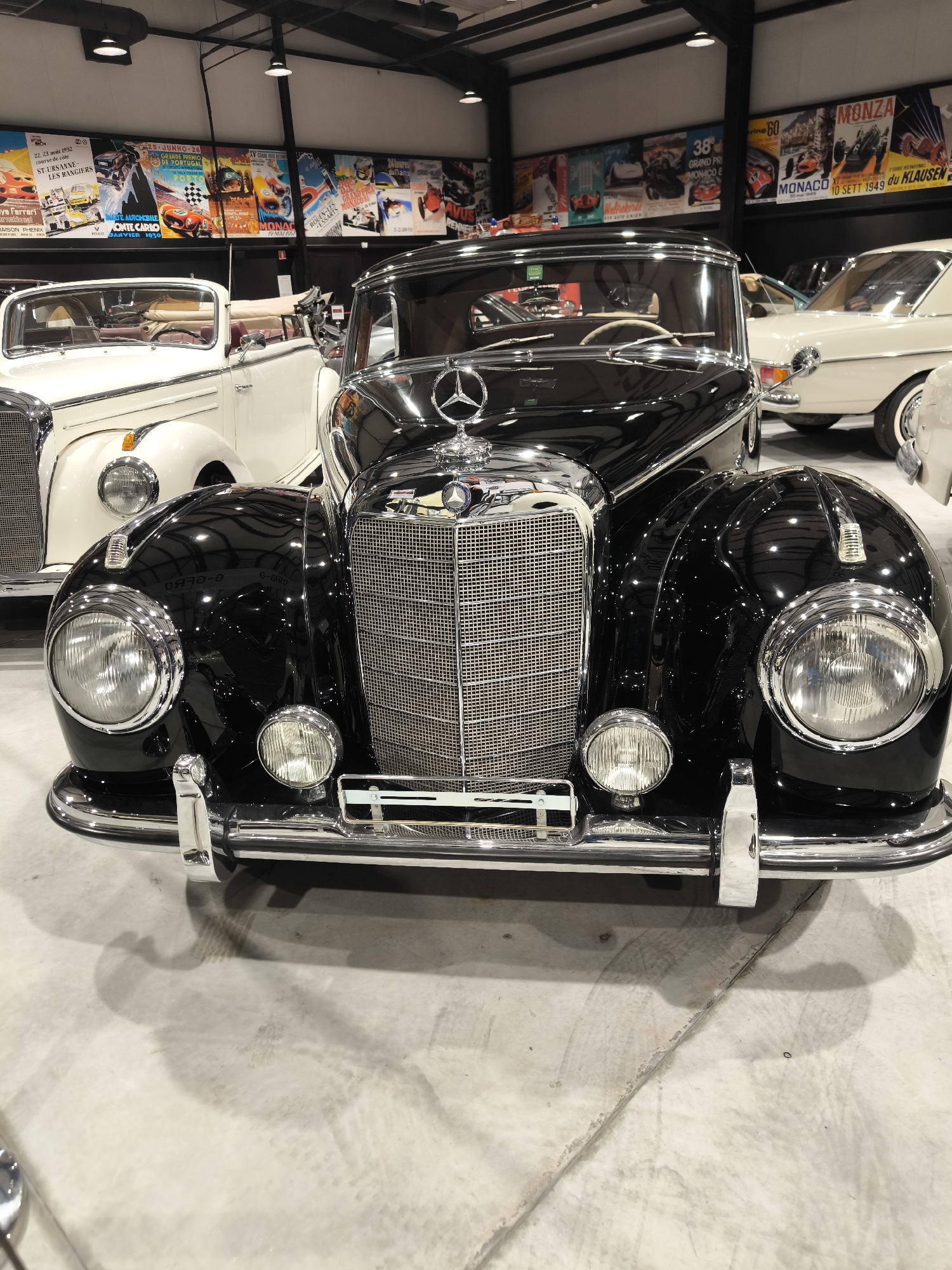 The image shows a close-up, frontal view of a classic black Mercedes-Benz, likely from the 1950s, with a prominent chrome grill and hood ornament. The car is surrounded by other vintage vehicles, including a white convertible Mercedes-Benz, inside what appears to be a museum or showroom featuring vintage racing posters along the back wall. The location in Kapatovo, Bulgaria is not evident from the visual details.