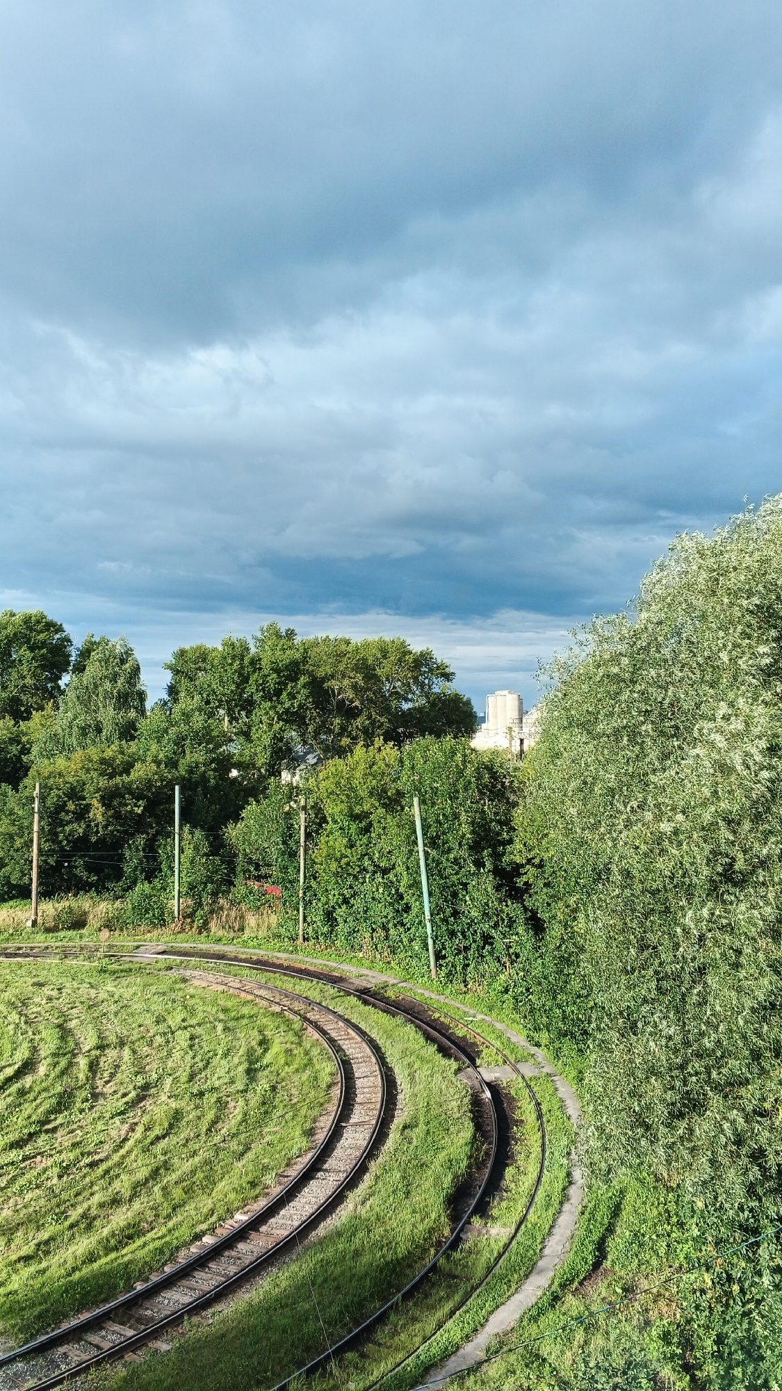 This image depicts a scene with railway tracks curving through a grassy area, bordered by lush green trees. In the background, industrial buildings are visible under a dramatic sky filled with clouds. The perspective is from an elevated viewpoint, looking down and across the landscape. The image does not contain any people or specific actions. Based on the provided context, this scene is located in the city of Izhevsk, Russia.
