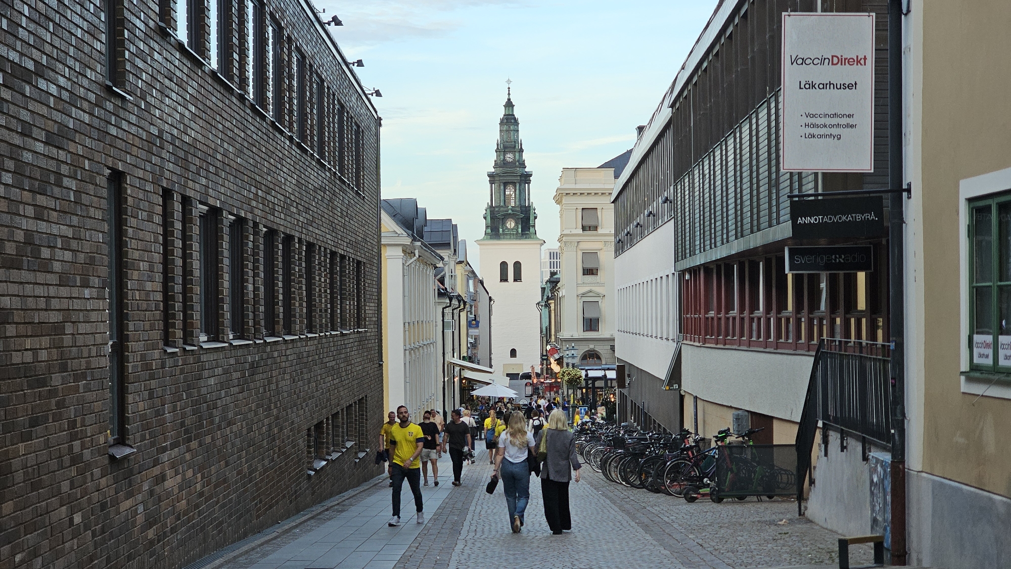 The image depicts a street scene in Linköping, Sweden, looking towards a church tower. On the left, a brick building with multiple windows runs along the street. On the right, a modern building with a slatted facade is visible, featuring signs for "VaccinDirekt Läkarhuset," "ANNTAdvokatbyrå," and "Sveriges Radio." A clock tower with a green spire is prominent in the distance, topped with a cross. Several people are walking down the cobblestone street, some are in groups, and others are walking alone. Bicycles are parked along the right side of the street. The sky is overcast, and the lighting suggests it is daytime.