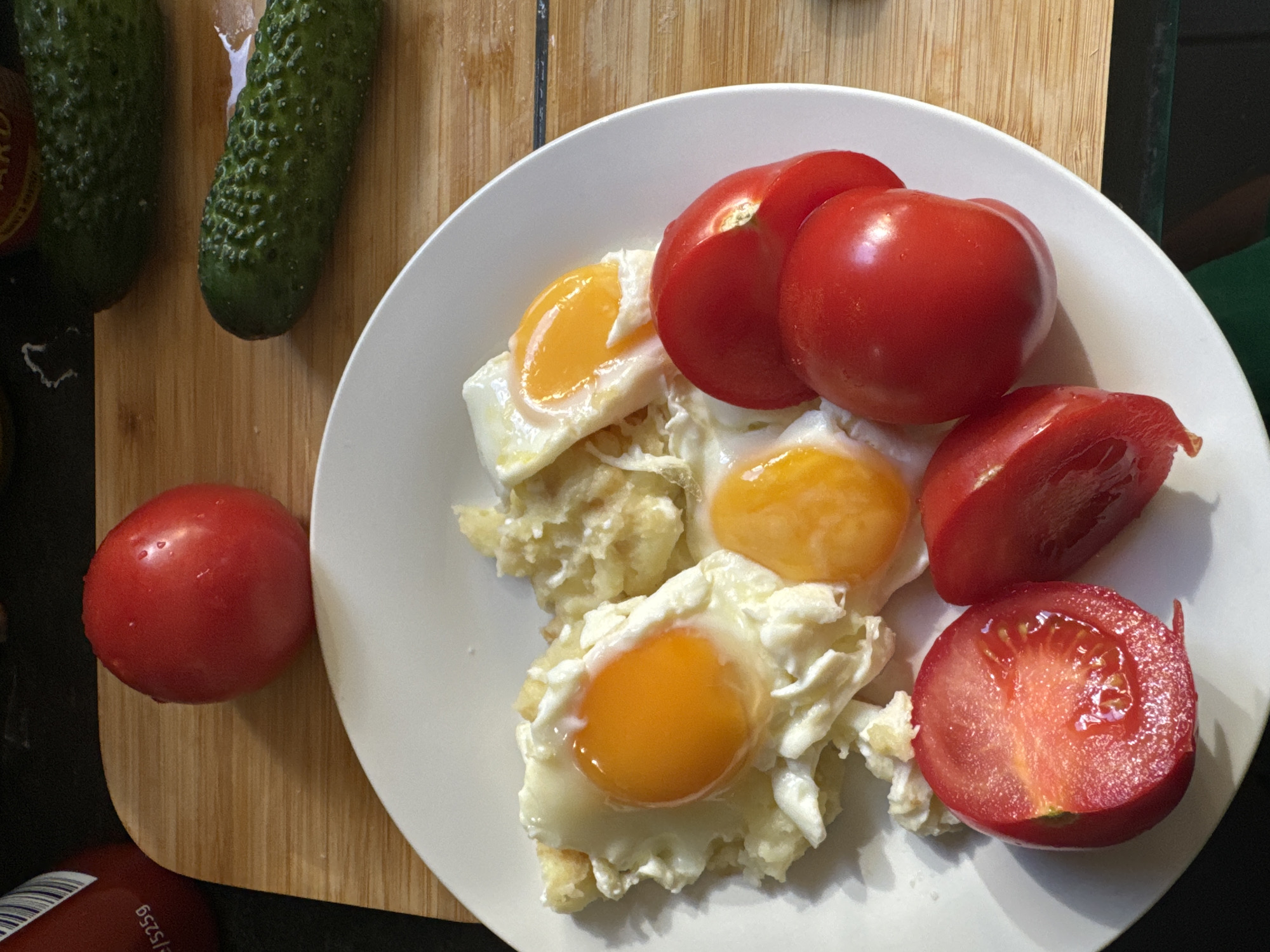 A plate of fried eggs and sliced tomatoes is presented on a wooden surface, alongside two whole cucumbers and a partially visible bottle of ketchup. The scene appears to be a domestic setting, likely a kitchen counter. There is no indication of the city of Waterford, Ireland. The lighting suggests it is daytime, with sunlight casting shadows. The food is prepared and ready to be consumed. The overall impression is one of a simple, homemade meal.
