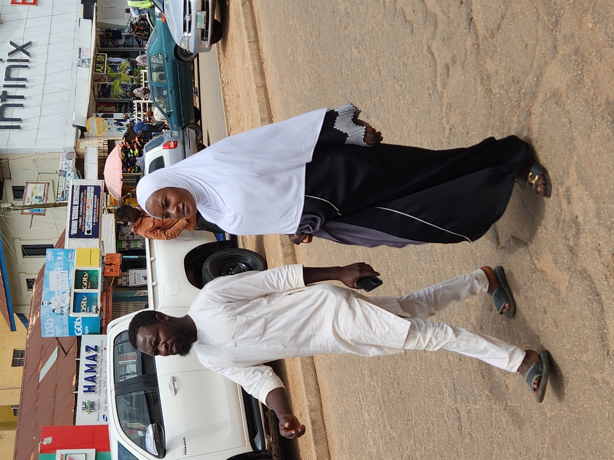 This image captures a street scene in Jos, Nigeria. In the foreground, a man and a woman are walking. The man, on the left, is wearing a traditional white tunic and trousers, with a black beard. He is holding a dark-colored mobile phone in his right hand and wearing dark sandals. The woman, on the right, is wearing a white hijab and a black and grey garment, also wearing sandals. They are walking on a paved road with visible cracks and patches.
In the background, there are various buildings and vehicles. To the left, a white pickup truck is parked, with a sign above it that reads "HAMAZ". Further back, a building with a red roof has multiple signs, including one for "DSB Nigeria FOOTBALL". Another building features a prominent "Infinix" logo. Several other vehicles, including a green sedan and a silver car, are visible on the road. The environment suggests a commercial area with shops and ongoing urban activity. The lighting indicates it is daytime, likely under direct sunlight.