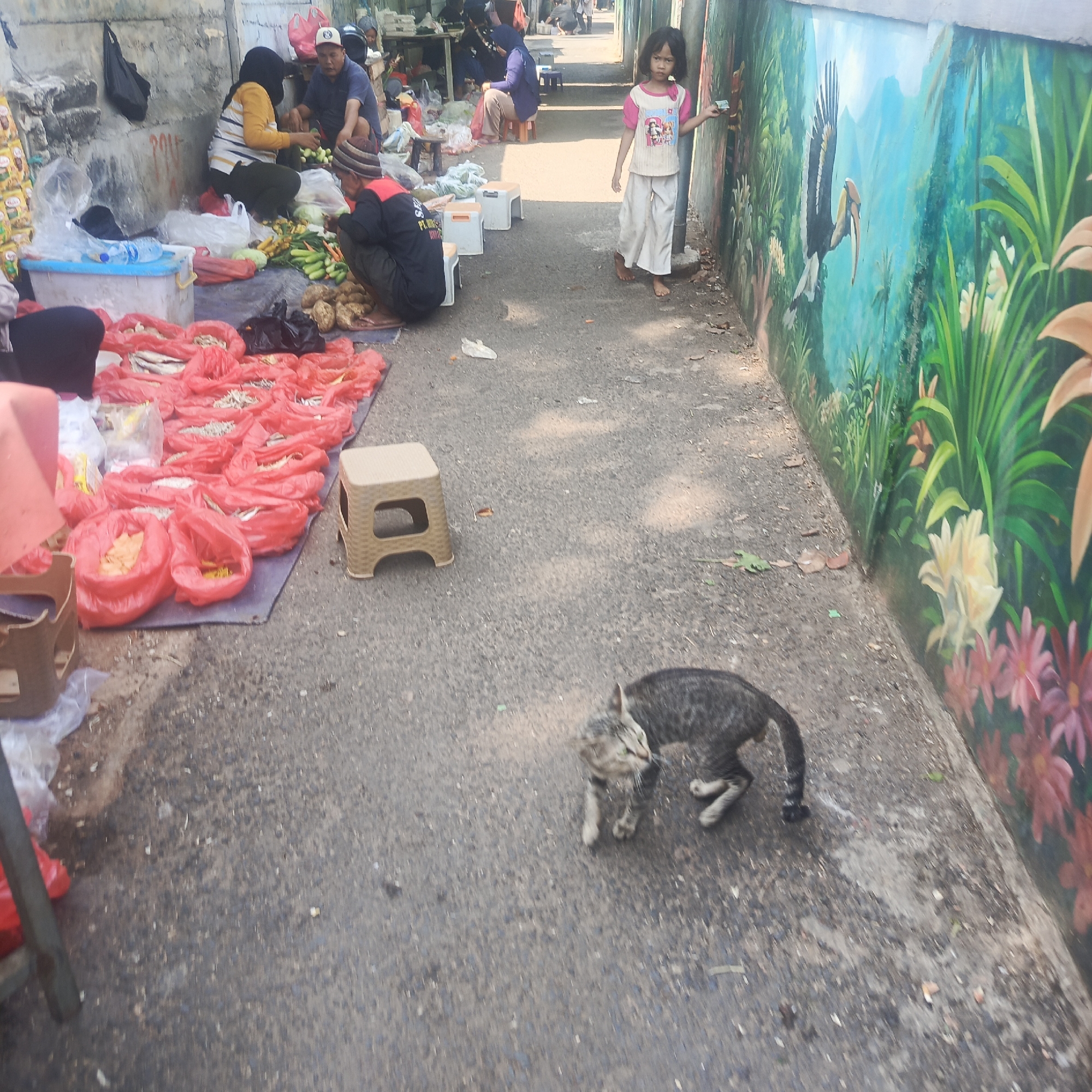 In South Jakarta, Indonesia, an outdoor market scene unfolds. The foreground features a tabby cat walking on an asphalt surface. The market stall area, to the left, displays an array of goods, primarily packaged in red plastic bags, alongside a blue plastic tub and a beige plastic stool. Several individuals are present at the stalls, including a woman in a yellow and white striped shirt and headscarf, a man in a dark blue shirt and cap, and another person in a dark shirt with a red trim. They appear to be engaged in market activities. Further back, a young girl in a pink and white shirt and white pants stands near a wall adorned with a vibrant mural depicting a tropical landscape with a bird. The overall environment suggests a bustling marketplace in an urban setting.