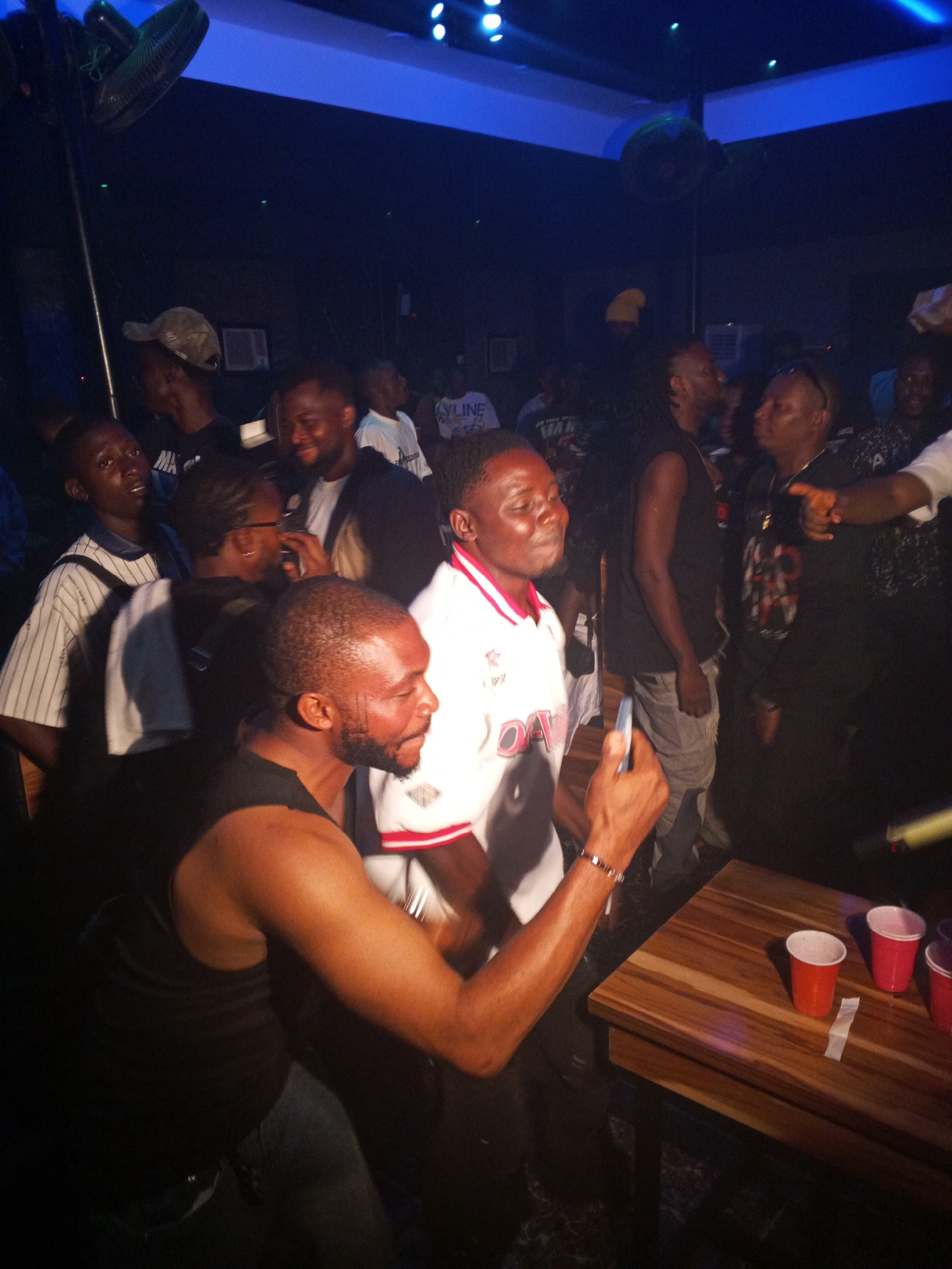 A group of people are gathered in a dimly lit indoor space, possibly a nightclub, in Lokoja, Nigeria. Men in the foreground are engaged in taking a selfie with a mobile phone. Other individuals stand around, some looking towards the camera while others are in conversation. A wooden table to the right holds several red plastic cups. In the background, fans are visible on the ceiling, and there is ambient lighting on the ceiling.