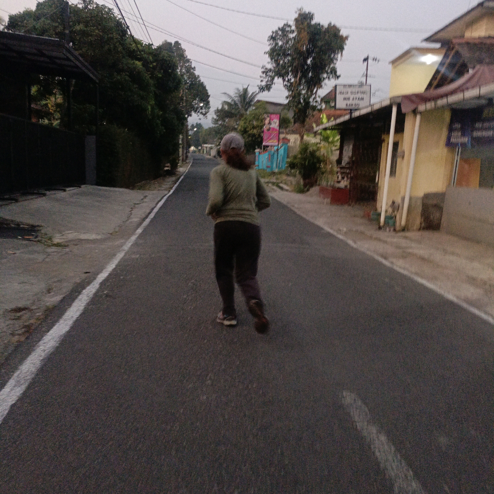 The image depicts a person from behind, wearing a light green long-sleeved shirt and dark pants, jogging on a paved road. They are also wearing a light-colored cap and athletic shoes. The road has a white line marking the edge on the left side. The surroundings suggest a semi-urban or rural environment in Salatiga, Indonesia, with trees, vegetation, and buildings visible on both sides of the road. On the right side, there is a building with a yellow facade and a brown roof, and a sign with text that appears to be in Indonesian, reading "NASI GORENG" and "MIE AYAM BAKSO". The sky is overcast, indicating it might be dawn or dusk. The overall scene conveys a sense of everyday activity in a local Indonesian setting.