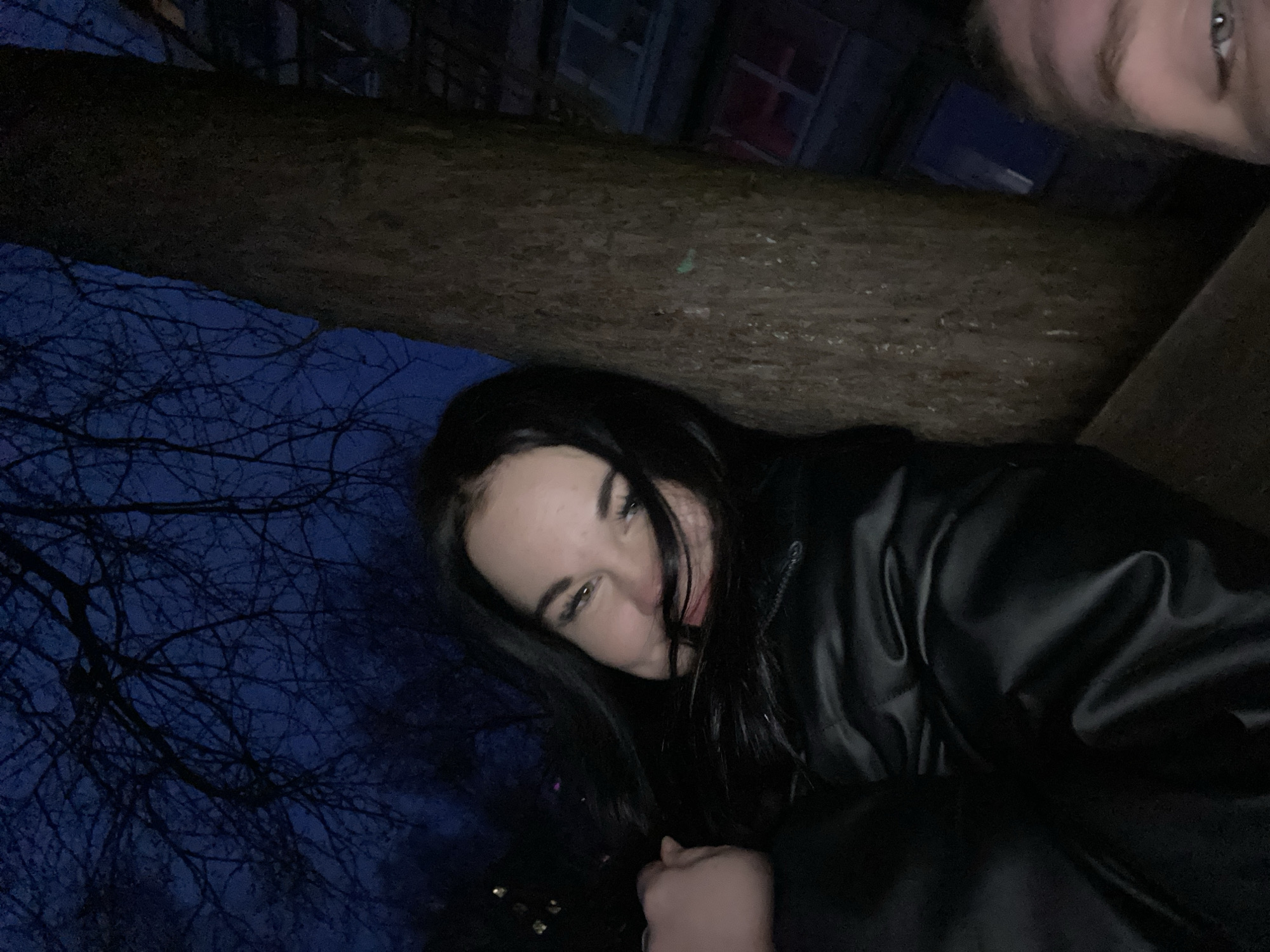 A low-angle, horizontally-oriented photograph depicts two individuals outside in a dimly lit environment. A woman with long, dark hair and fair skin is positioned centrally. She wears a dark leather jacket and looks downward with a slight smile. To her upper right, partially cut off, is another person's face, with one eye visible. Above the woman is what appears to be a thick tree branch, partially obscuring a building in the background. Bare tree branches are silhouetted against a dark blue sky to the left. Based on the prompt, the location is Chaikovsky, Russia.