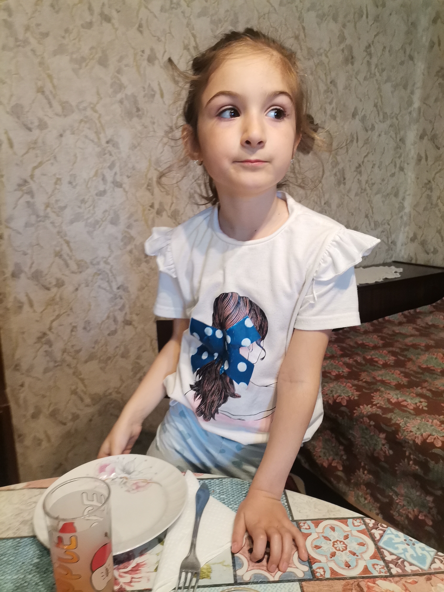 A young girl with light brown hair styled in a loose updo stands at a table. She is wearing a white t-shirt with short, ruffled sleeves and a graphic of a girl with a large blue polka-dot bow. The girl is looking to her left with a slightly pensive expression.
On the table in front of her is a plate, a glass of liquid (possibly juice), a napkin, and a fork. The table has a colorful tiled pattern. Behind the girl is a wall with a light-colored, mottled wallpaper and a bed with a floral patterned cover. The location is likely an indoor dining or kitchen area in a residential setting. The city of Pobeda, Bulgaria is not verifiable from the image content alone.