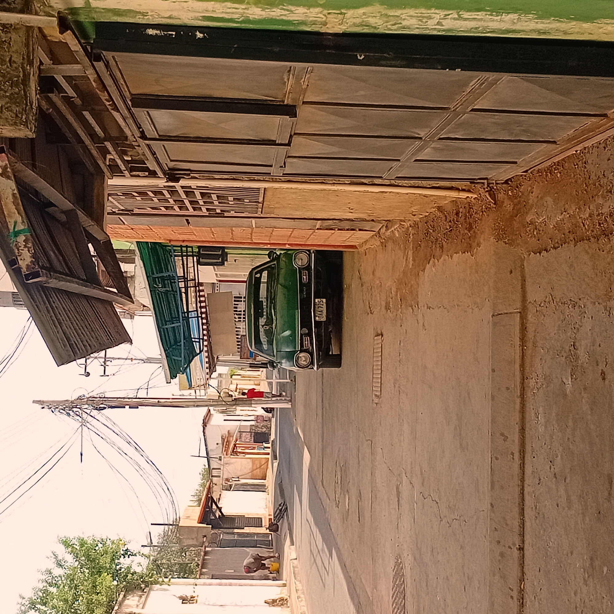The image depicts a narrow street in an urban area. A green car is parked on the street. Overhead electrical wires are visible. A person is working near a building, appearing to be painting or washing something. The buildings have a mix of materials and styles, including metal, brick, and stucco. The environment is dusty and weathered.