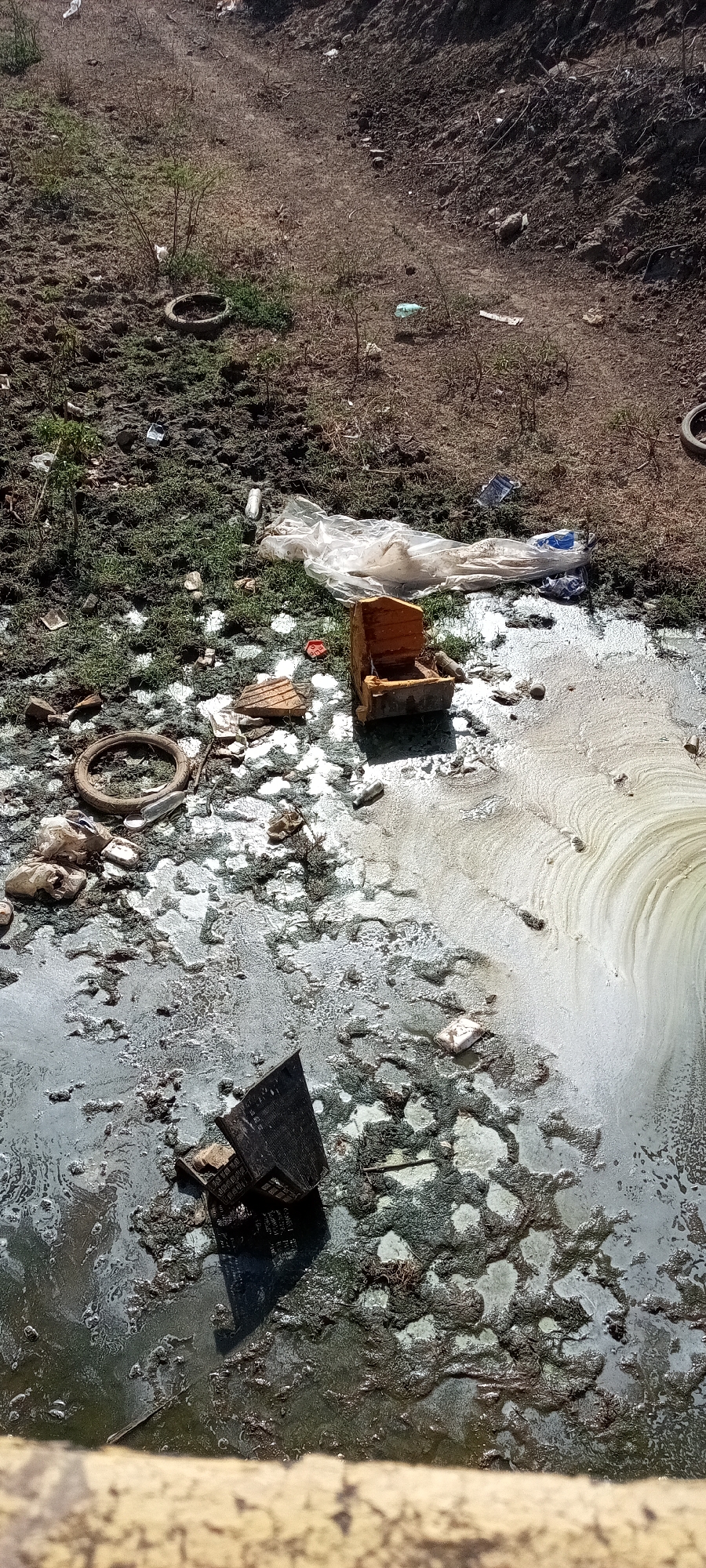 The image depicts a polluted, presumably urban, environment. The location appears to be a ditch or gully filled with stagnant water, possibly wastewater. The water surface is covered with a layer of scum and what appears to be soap suds or foam, suggesting chemical contamination. A variety of refuse and debris is visible both in the water and on the adjacent bank. These include old tires, pieces of wood furniture, plastic sheeting, and other unidentifiable garbage. The bank is sparsely vegetated and mostly barren soil. The location is likely in the city of Unknown, Unknown based on the request.