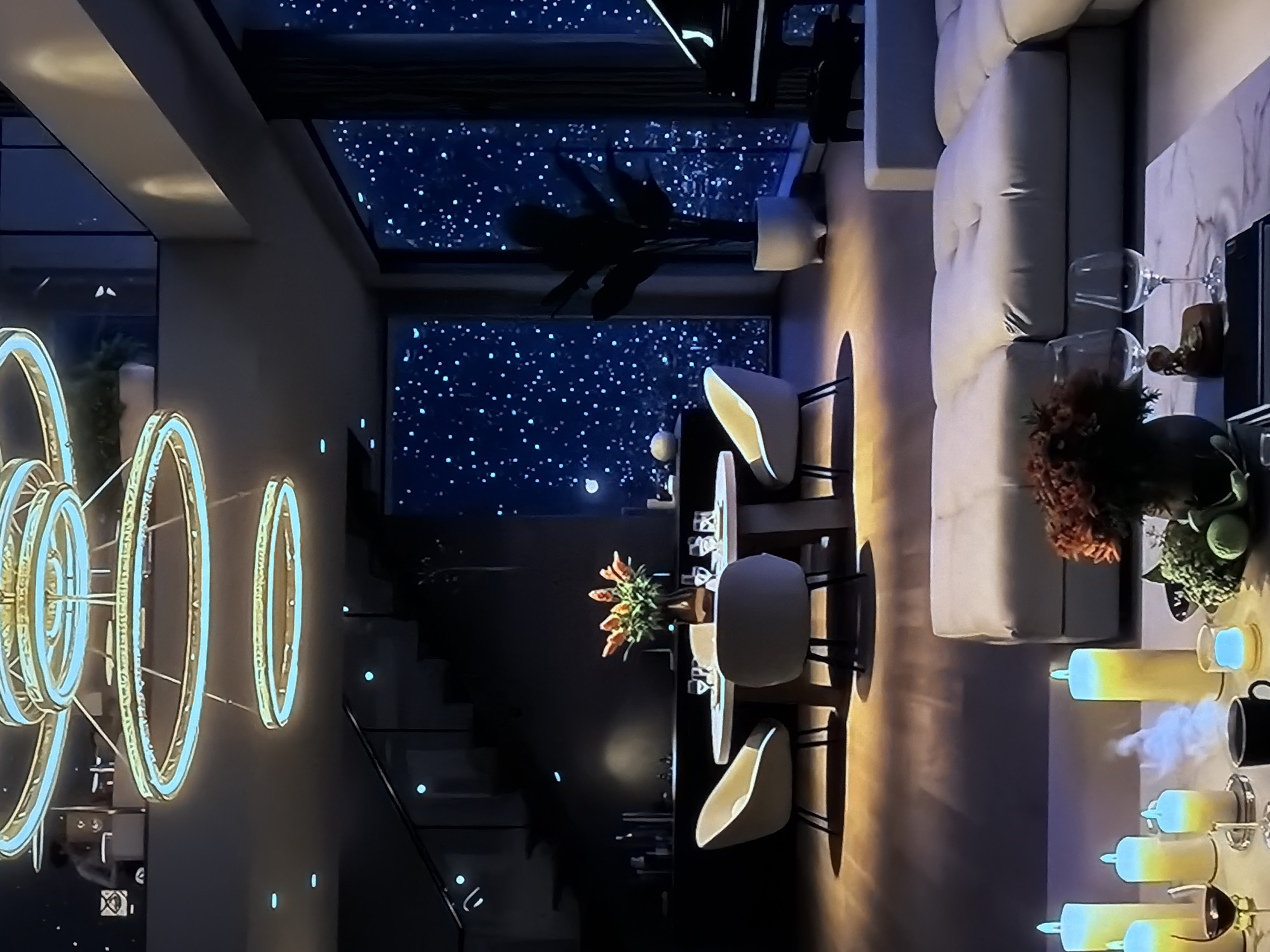 This media file captures a modern, luxurious interior space, likely a living and dining area, at night in Sofiivska Borshchahivka, Ukraine. No people are visible.
The scene features a spacious living room on the right, furnished with a large, light-colored L-shaped sofa. A marble-topped coffee table sits in front of it, displaying two wine glasses, a small decorative figure, and some dark foliage. Adjacent to the sofa, several unique decorative candles or lights glow with a transition from yellow to blue, some emitting a misty vapor. A black mug is also on this surface.
To the left of the living area is a dining space, marked by a table with four elegant chairs with light seats and dark bases. A vase of vibrant orange flowers serves as a centerpiece on the dining table. Beyond this, a grand, multi-ringed chandelier with a light blue glow hangs in a darker, potentially double-height space.
The most striking feature is the large windows that line the back wall, revealing a deep blue "sky" filled with countless tiny, bright points of light, creating the illusion of a starry night. A silhouetted plant is visible against one of these windows. In the background, a modern staircase with glass railings features subtle blue accent lighting on its steps. The overall lighting is dim and atmospheric, with warm glows from various fixtures complementing the cool blue of the windows and some decorative elements. The setting evokes a serene, sophisticated, and possibly romantic ambiance. No visible text is present in the image.