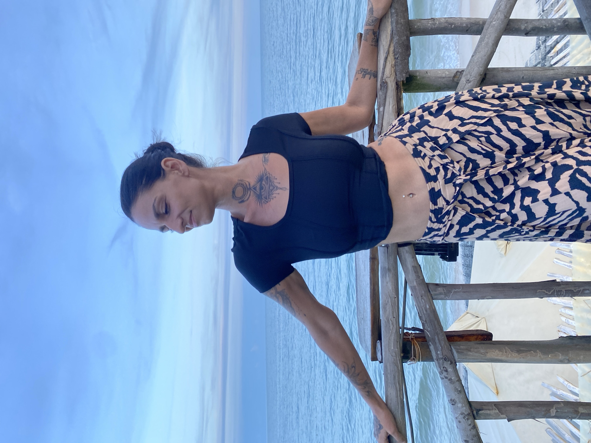 A medium shot captures a woman standing on a wooden walkway overlooking the ocean at dusk. She has her hair pulled up in a bun and is wearing a black crop top and a patterned skirt. She has several tattoos, including on her neck, chest, and arms. The ocean is calm, and the sky is a soft blue with hints of pink and orange. In the background, there appears to be a beach with some structures, possibly tents or cabanas. The overall impression is one of peacefulness and tranquility.
