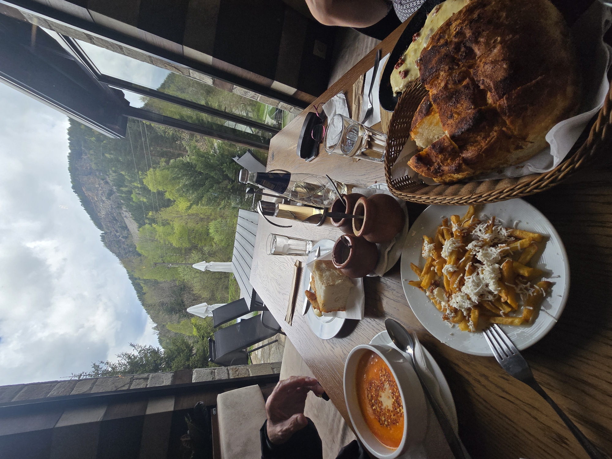 The image captures a dining scene at a restaurant, potentially in Sapareva Banya, Bulgaria, given the mountainous landscape visible through the window. The table is set with various dishes: a basket of bread, a plate of french fries topped with cheese, a bowl of soup, and small clay pots. Cutlery is laid out, and drinks, including a bottle and glasses, are present. An individual is visible in the foreground, and another person is partially visible in the background. The setting appears to be indoors with a window view overlooking a mountain landscape, suggesting a restaurant location.