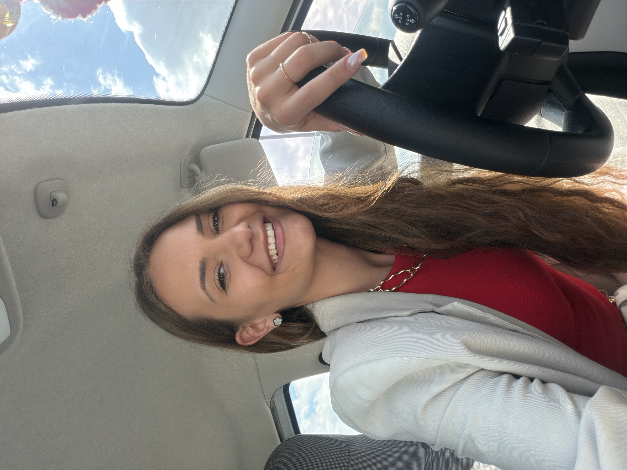 A young adult female is depicted seated in the driver's side of a vehicle in Sofia, Bulgaria. She has long, wavy brown hair, fair skin, and dark eyes, looking directly at the camera with a wide smile revealing her teeth. She wears a light-colored blazer over a red top, adorned with a gold-colored chain necklace and small, white, flower-shaped stud earrings. Her left hand, featuring a light-colored manicure with yellowed tips and two thin gold-colored rings on her ring and middle fingers, grips the top right portion of a black steering wheel. A control button cluster with directional arrows is visible on the steering wheel's right spoke. The vehicle's interior features a light grey fabric ceiling. A sunroof on the right side of the frame reveals a bright blue sky with white clouds and the partial presence of what appear to be reflective balloons. Sunlight illuminates the scene from above. A portion of a grey fabric car seat is visible on the left.