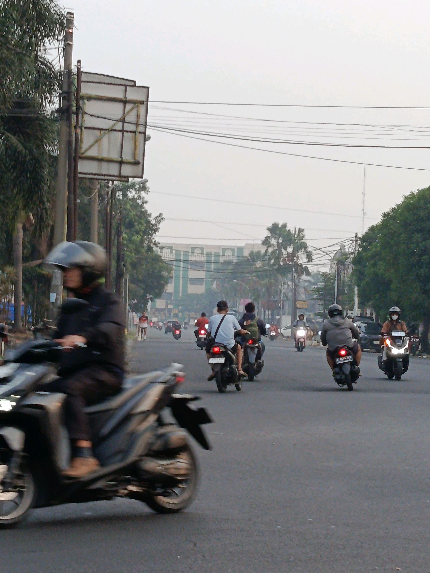 The image depicts a street scene in Bekasi, Indonesia, during what appears to be the early morning or late afternoon given the subdued lighting and overcast sky. The road is filled with numerous motorcycles, indicating it's a common mode of transportation in the area.
In the foreground, a person wearing a black jacket and helmet rides a dark-colored scooter, their body angled slightly to the left. The scooter's front wheel is in motion, suggesting the rider is actively moving. Behind this, several other motorcycles are visible, each with riders. Some are close-up, showing details of their clothing and helmets, while others are further away, appearing as smaller forms on the road.
The environment features a paved road with white lane markings. On the left side of the road, there is a tall pole with a large, blank billboard structure attached to it, surrounded by palm trees. Further down the road, a pedestrian wearing a red shirt and dark shorts is walking. The right side of the road is lined with trees and a building with a distinct architectural style, featuring arched windows and a pale green facade, suggesting a commercial or institutional structure. Overhead, power lines crisscross the sky. The overall atmosphere is one of a busy urban thoroughfare with moderate traffic.