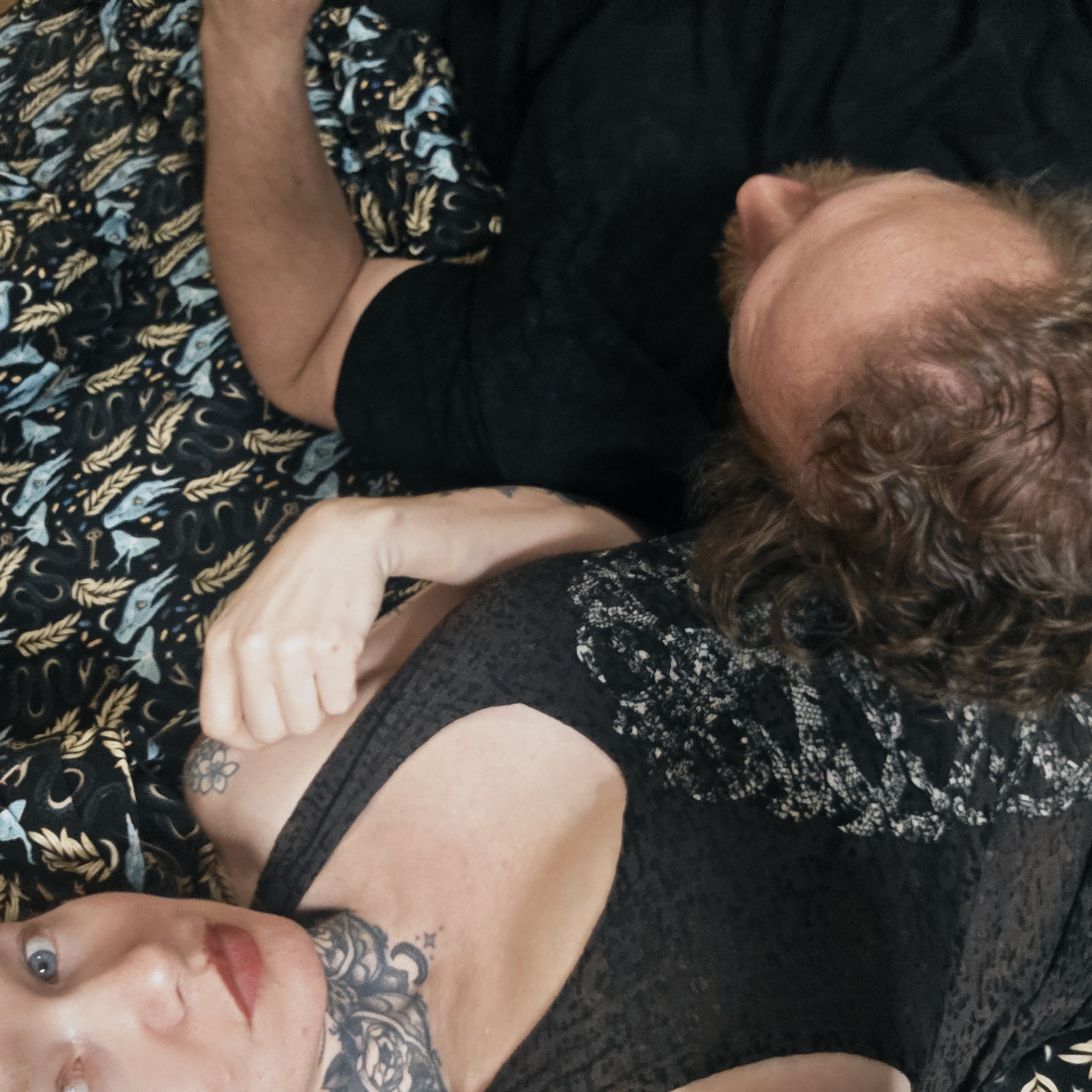 Two people are lying on a bed with a patterned bedspread of black, gold and blue. The person closest to the camera is a woman with light skin, blue eyes and red lipstick. She has a prominent tattoo on her neck and another tattoo visible on her upper chest. Her left arm is bent and her fist is near her chest. She is wearing a dark, semi-sheer top. Behind her is a man with light skin and brown hair, wearing a black shirt. He is looking up. The location cannot be determined to be in Saint Petersburg, Russia.