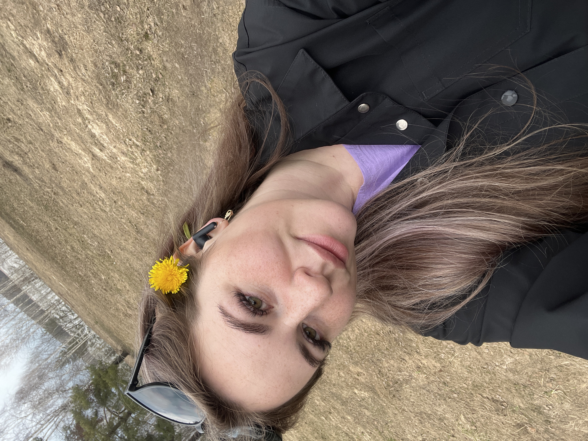 A woman with fair skin, freckles, and light brown hair is lying on a grassy surface, likely outdoors. She has a dandelion flower tucked behind her ear and sunglasses resting on her hair. She's wearing a black jacket and a purple shirt. The background appears to be a field or open grassy area with trees visible in the distance. The photo may have been taken in a park, given the context of Chaikovsky, Russia.
