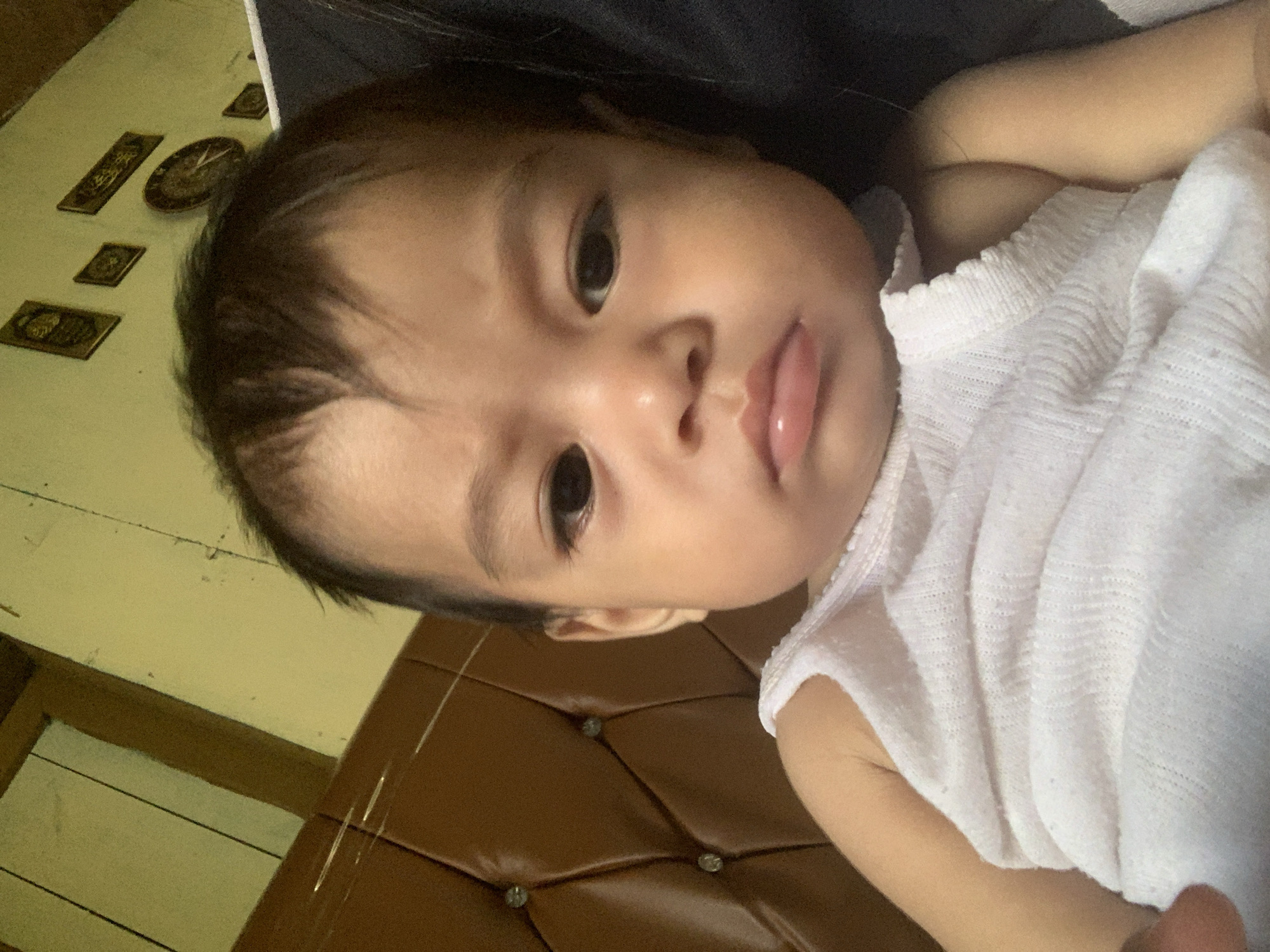 A close-up, frontal, slightly-above-eye-level shot shows a young infant with dark hair, dark eyes, and chubby cheeks. The infant is wearing a white, ribbed tank top. The background is a brown, tufted surface on the left and a wall with several framed pictures and a clock on the right. The lighting appears to be from above and slightly to the left, casting a slight shadow. The overall scene suggests an indoor setting, possibly a home environment. There is no information in the image to determine the specific location within Sungai Meriam, Indonesia.