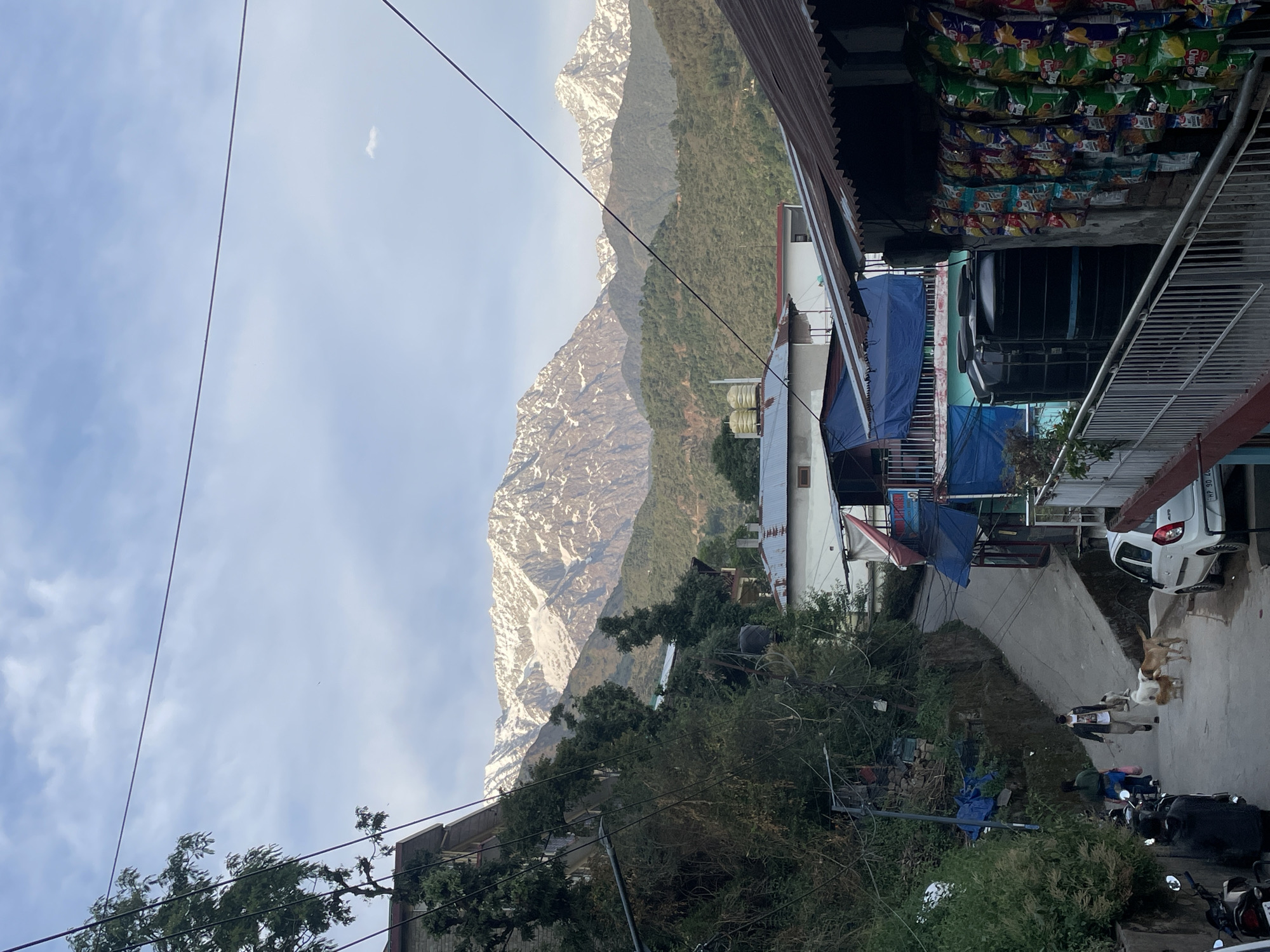 The scene is an outdoor view in Dharamshala, India, featuring a mountainous backdrop partially covered with snow. The image is taken from an elevated position, overlooking a street with several buildings and structures. Overhead power lines intersect the sky. A store is visible with snack items stacked on shelves, and beneath it, a blue tarp partially covers an area.
Below, a paved road slopes down, with parked motorcycles and a white car. Several dogs are on the street along with a person walking. Vegetation includes trees and shrubs. The sky is overcast with a few clouds.