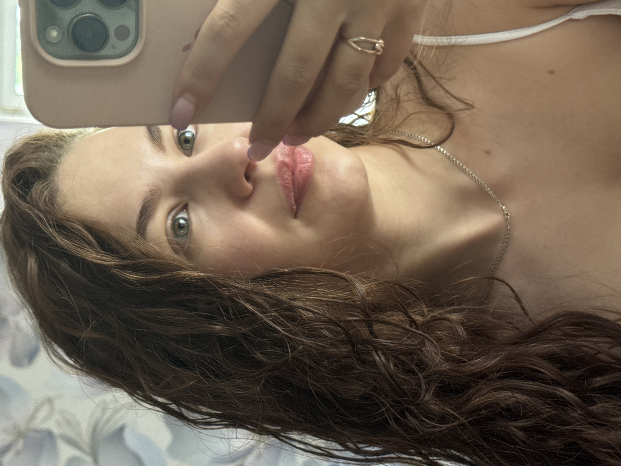 A woman takes a selfie with a phone. The background appears to be a patterned surface, possibly bedding. The woman has brown, curly hair and green eyes. She is wearing a silver necklace and a gold ring. The phone case is light pink.