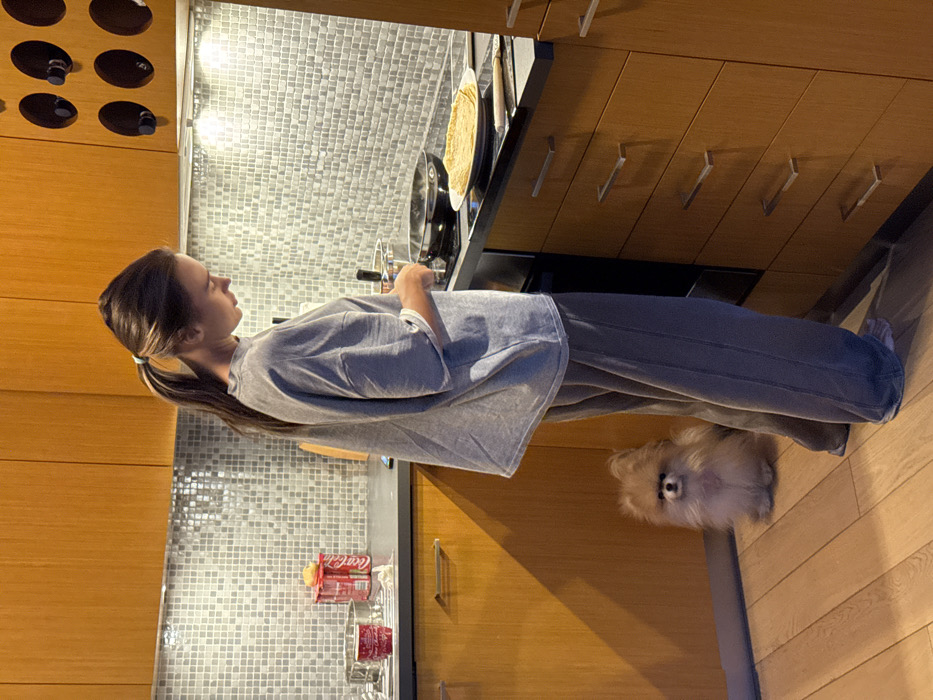 An adult female is depicted standing in a modern kitchen, presumably located in Warsaw, Poland. She is dressed in a light grey oversized t-shirt and dark grey sweatpants, with her brown hair tied back in a ponytail. Her gaze is directed downward, focused on a black frying pan on an induction or electric stovetop, from which steam is visible. She appears to be actively cooking, holding a utensil in her right hand over the pan.
To the right of the stovetop, a stack of freshly cooked crepes or pancakes rests on a white plate. The kitchen features light wooden cabinetry with horizontal brushed metal handles and a grey mosaic tile backsplash. Under-cabinet lighting illuminates the workspace. On the far left of the black countertop, two red Coca-Cola cans, a red mug, and a red cup are visible.
A small, light brown Pomeranian-type dog sits on the light wooden floor to the left of the female, positioned behind her legs, facing forward and looking towards the camera. The dog appears attentive. The floor is made of light-colored wooden planks.
