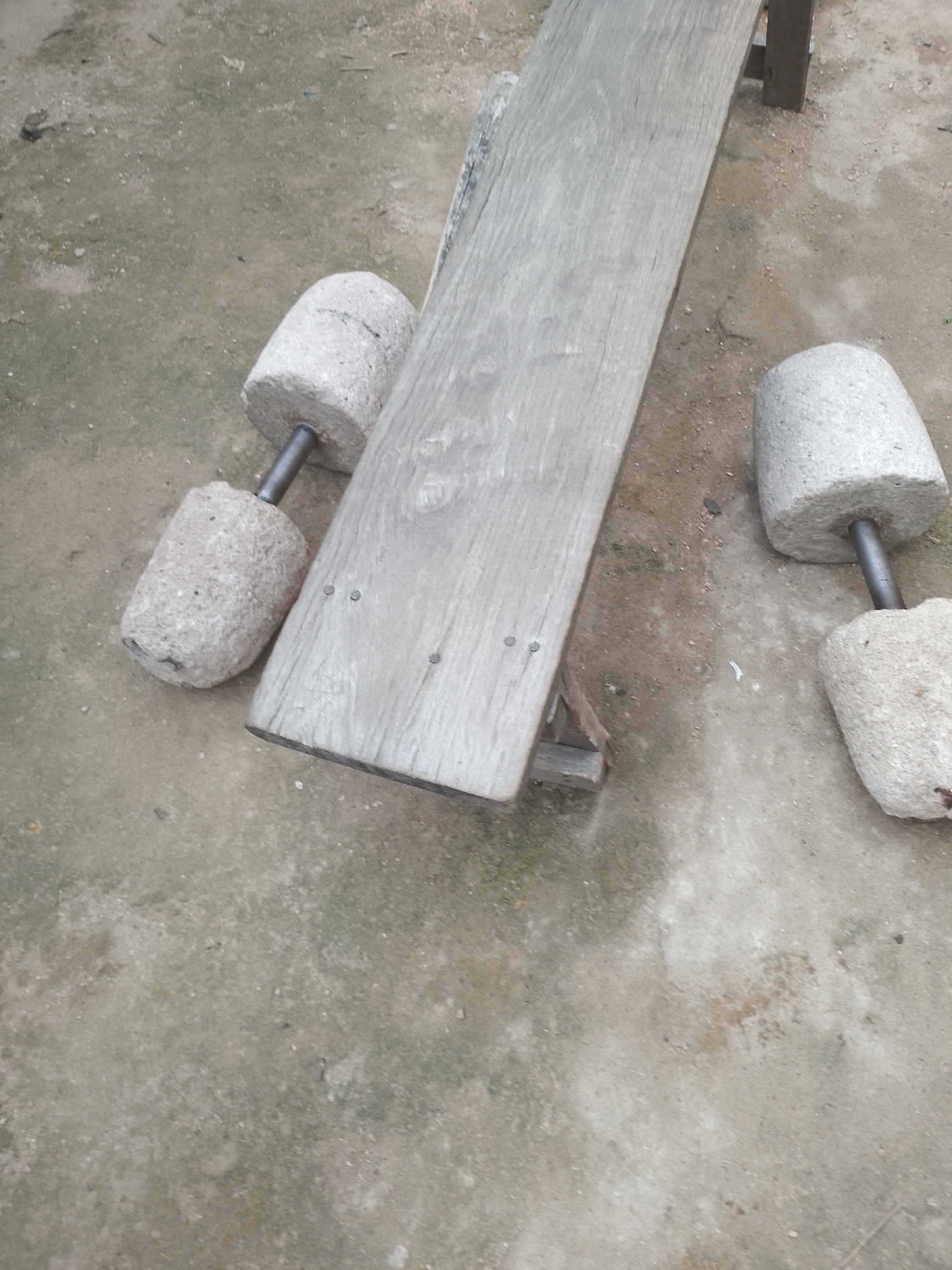 In an outdoor environment with a dirt and gravel ground, a weathered wooden plank rests on a simple wooden support structure. To the left of the plank, two cylindrical, rough-textured stone weights are mounted on metal rods, positioned at an angle. To the right of the plank, another similar stone weight, also on a metal rod, is visible. The overall scene suggests an area used for manual labor or traditional grinding activities.