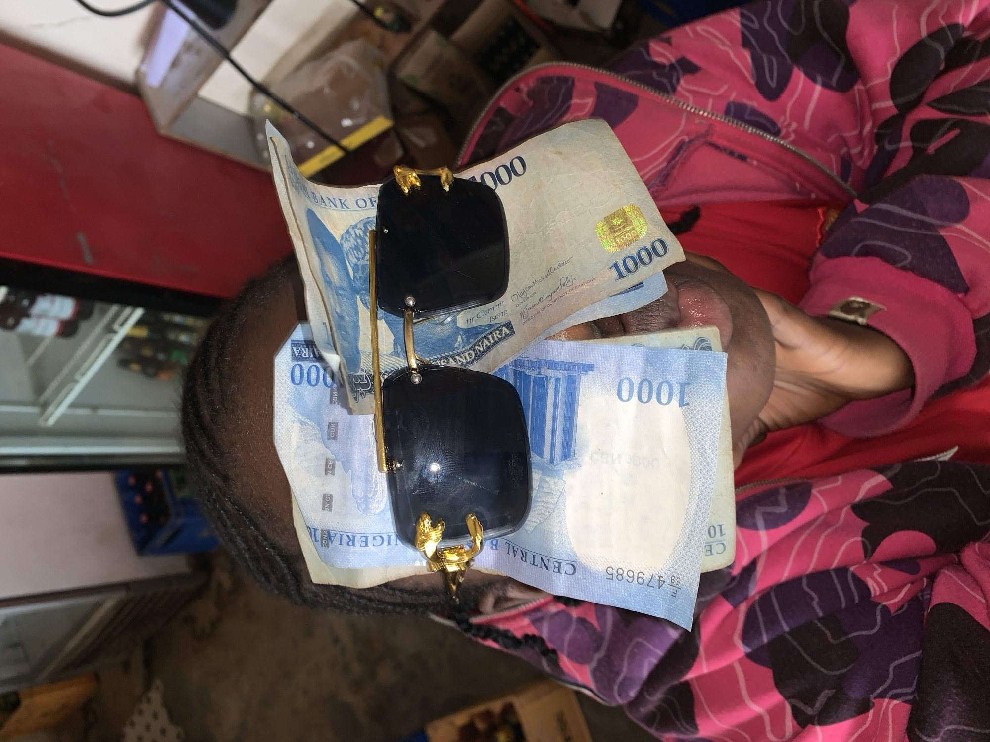 The image presents an overhead, close-up view of a person holding several Nigerian Naira 1000 banknotes, upon which a pair of dark sunglasses rests. The person is partially visible, showing their dark, braided hair on the left and their right hand holding the money on the right, wearing a jacket with a distinct pink and purple camouflage-like pattern.
The sunglasses have large, dark, rectangular lenses and prominent gold-colored frames, featuring intricate designs resembling animal heads or ornate clasps where the arms connect to the lenses. The banknotes are fanned out, clearly showing "1000" printed multiple times. Visible text on the currency includes "BANK OF," "NIGERIA," "CENTRAL BANK," the signatures of "Dr Clement Isong GOVERNOR" and "Oluyemi Michael Cardoso DIRECTOR OF CURRENCY OPERATIONS," and a serial number "F 59479685."
The background suggests an indoor commercial environment, possibly a shop or market stall, with a red surface, blurred bottles, and other items. The presence of Nigerian Naira banknotes strongly indicates the location is in Nigeria, specifically New Stadium as per context. The scene depicts a person holding or displaying a sum of money and sunglasses, possibly in preparation for a transaction or as a personal possession.