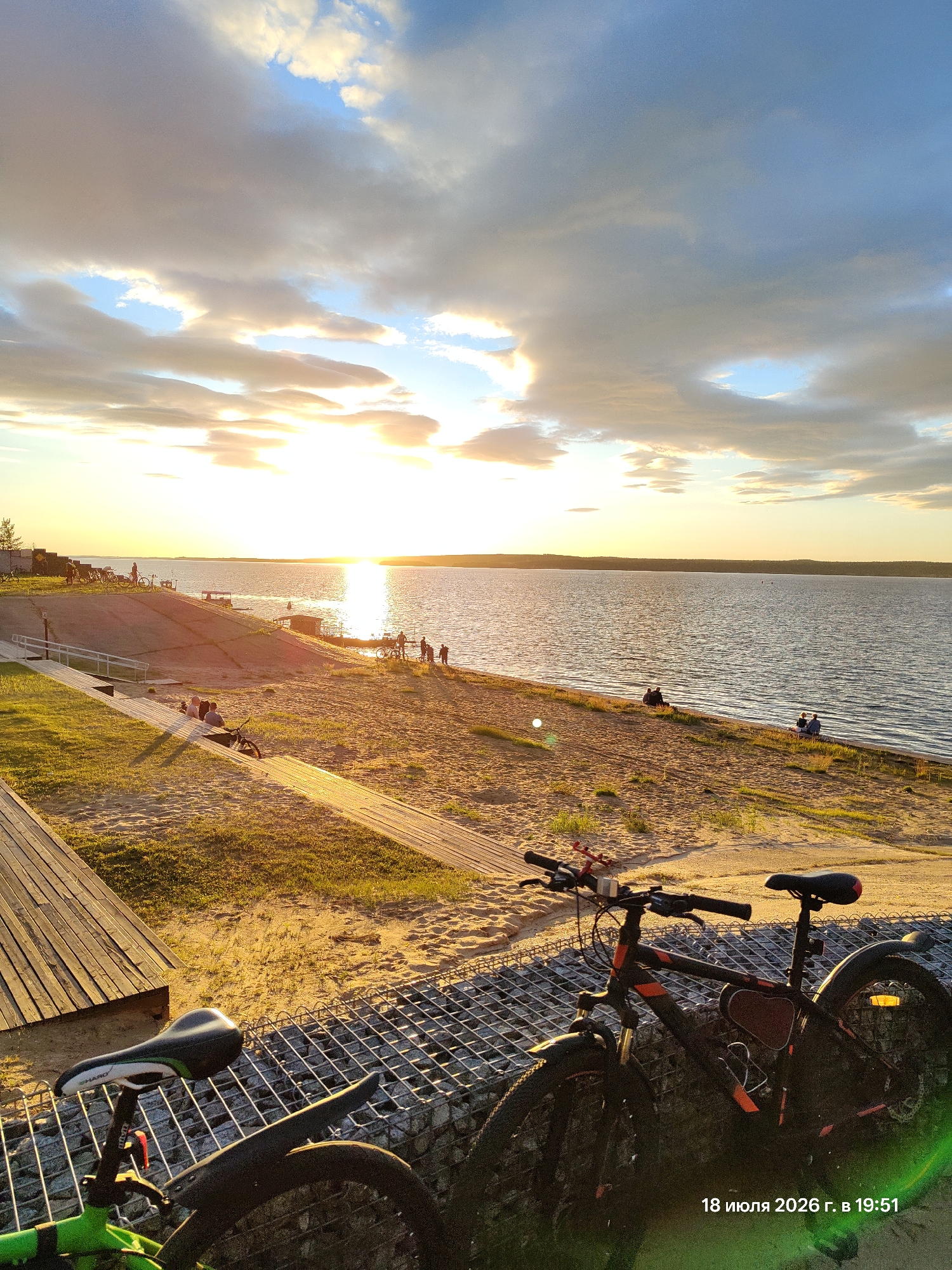 The image captures a sunset scene at a lakeside beach, possibly in an urban park. In the foreground, two bicycles are parked near a metal grate structure. One bicycle is green and partially visible on the left, while a black and orange bicycle is prominently displayed on the right. Behind the bicycles, a sandy beach stretches towards the water, with wooden pathways and grassy areas visible. Several people are scattered along the beach, some sitting and others standing, enjoying the view. The sun is low on the horizon, casting a warm glow across the water and the sky, which is filled with dramatic clouds. A timestamp "18 июля 2026 г. в 19:51" is visible in the lower right corner.