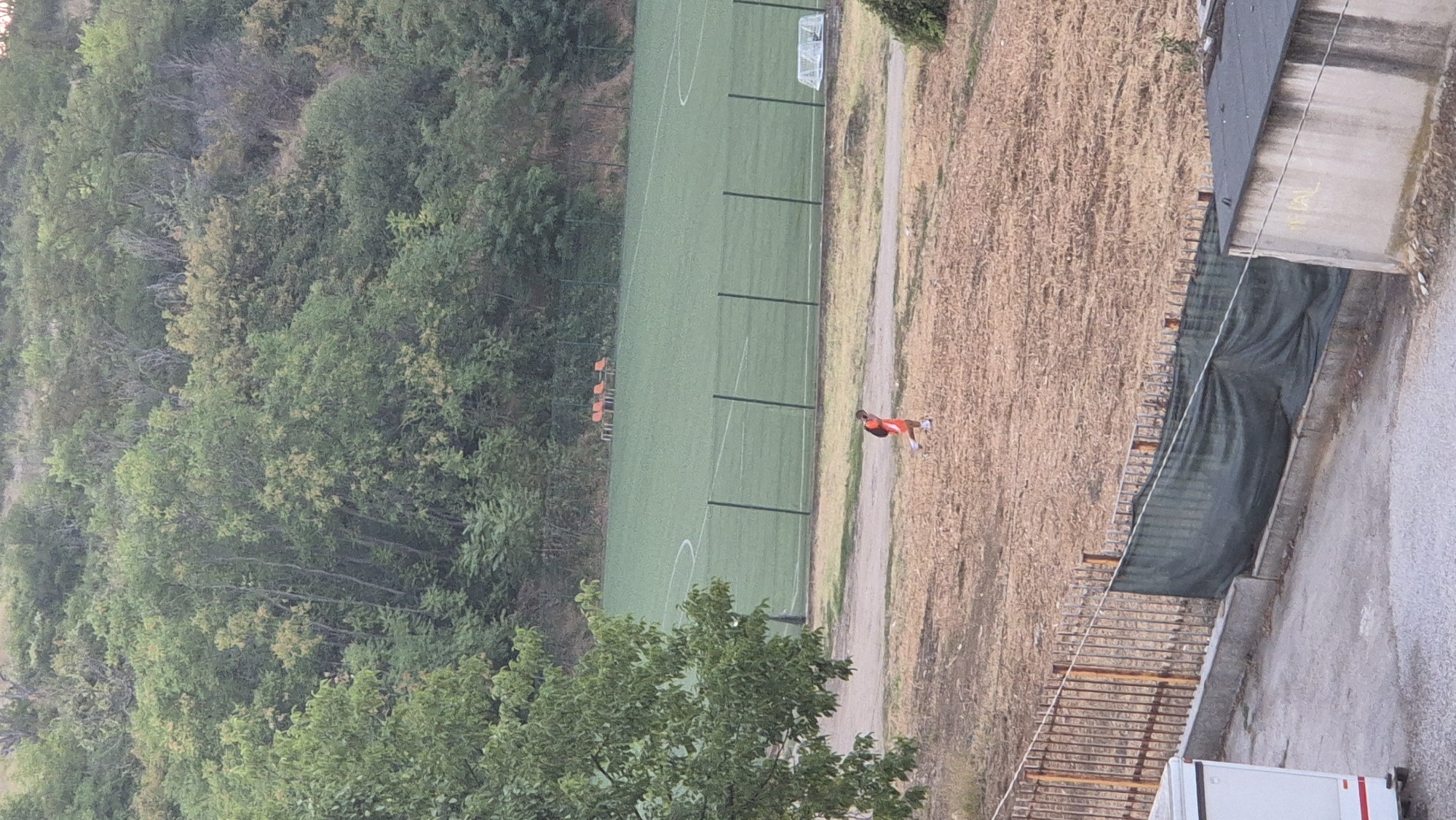 A person wearing an orange shirt and black shorts with an orange backpack is walking across a dry, grassy field. In the background, there is a green soccer field with white markings, a fence, and some orange seats. Behind the soccer field is a wooded hillside. In the foreground, there is a gray concrete wall with graffiti, a black fence with green netting, and part of a white trailer. The image appears to be taken from a higher vantage point, looking down on the scene. The location is not specified in the provided image or its crops.