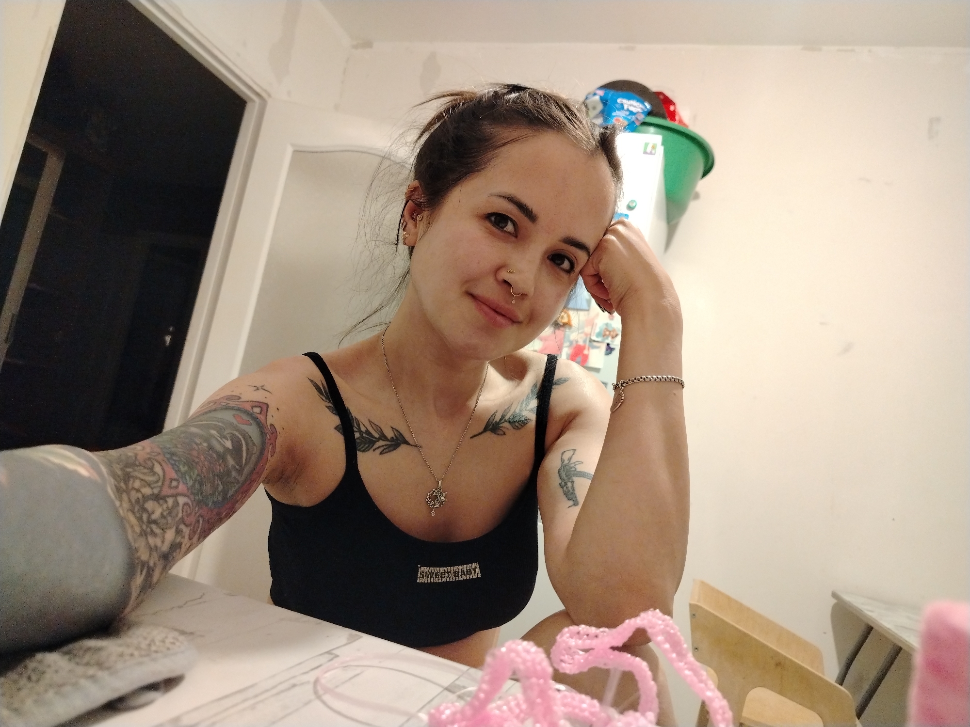 A selfie captures a woman in a black tank top, adorned with a necklace, wrist bracelet, and multiple tattoos on both arms and her chest. Her hair is pulled back, revealing a pierced nose and multiple ear piercings. She rests her face on her hand, smiling slightly. The setting appears to be indoors, with a doorway visible on the left. Behind her, a white wall is partially obscured by a green container and a white refrigerator. A table is in the foreground, with pink crafting materials. The specific location within Noviy, Russia, is not discernible from the image.