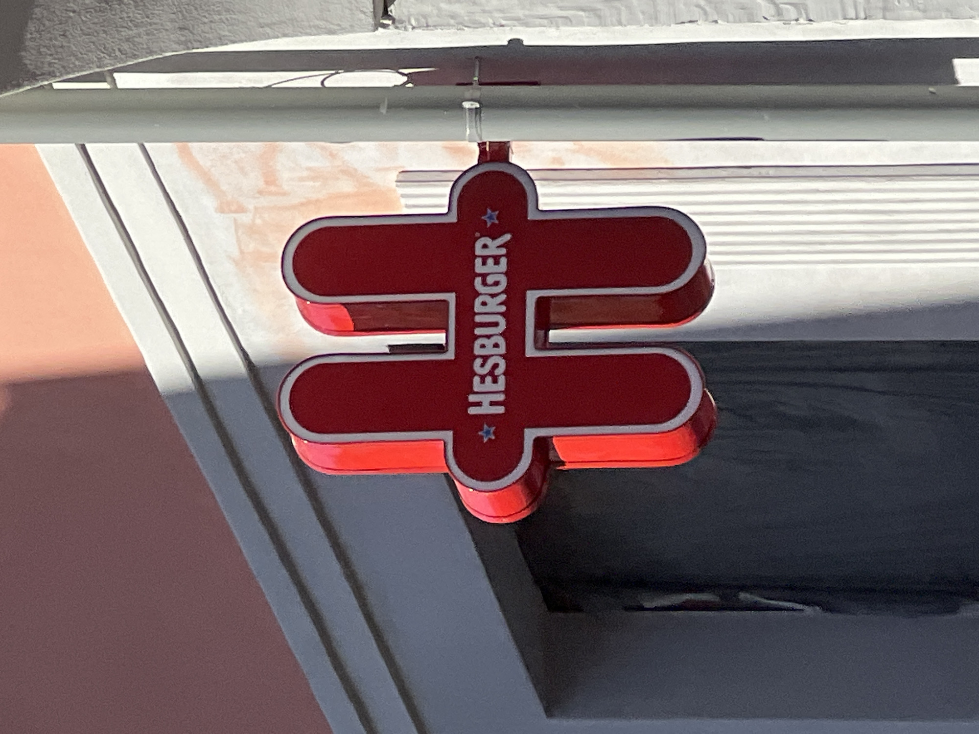A close-up shot captures a red "HESBURGER" sign suspended from a white pipe. The sign features a white outline around the red shape, which resembles an equals sign. The text "HESBURGER" is vertically oriented within the sign, flanked by stars. The background includes elements of the building's facade, revealing a pink wall, white trim, and a dark window. The setting appears to be an urban environment, likely near a Hesburger restaurant. The location is identified as Unknown, Unknown.