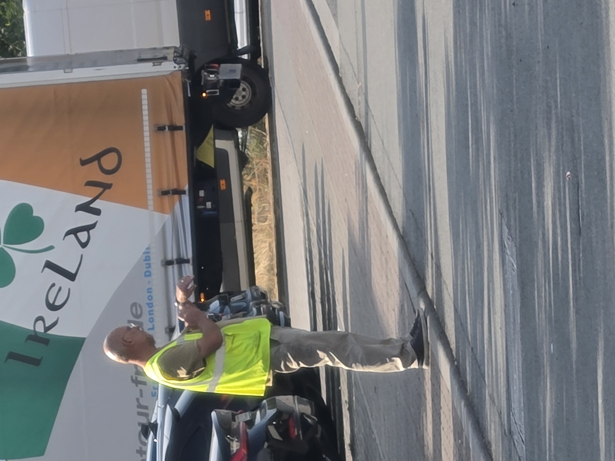 A bald, middle-aged man wearing a fluorescent yellow safety vest over a tan short-sleeved shirt and tan trousers stands on a sidewalk next to a parked dark car. He is looking at a watch on his left wrist. Behind him is a large white and orange truck with the word "Ireland" visible on its side, along with a green shamrock graphic. The truck has writing that appears to say "detour-fr" and "London-Dubl." The scene takes place outdoors on a sunny day, with shadows cast on the ground. This image was taken in Frankfurt, Germany.