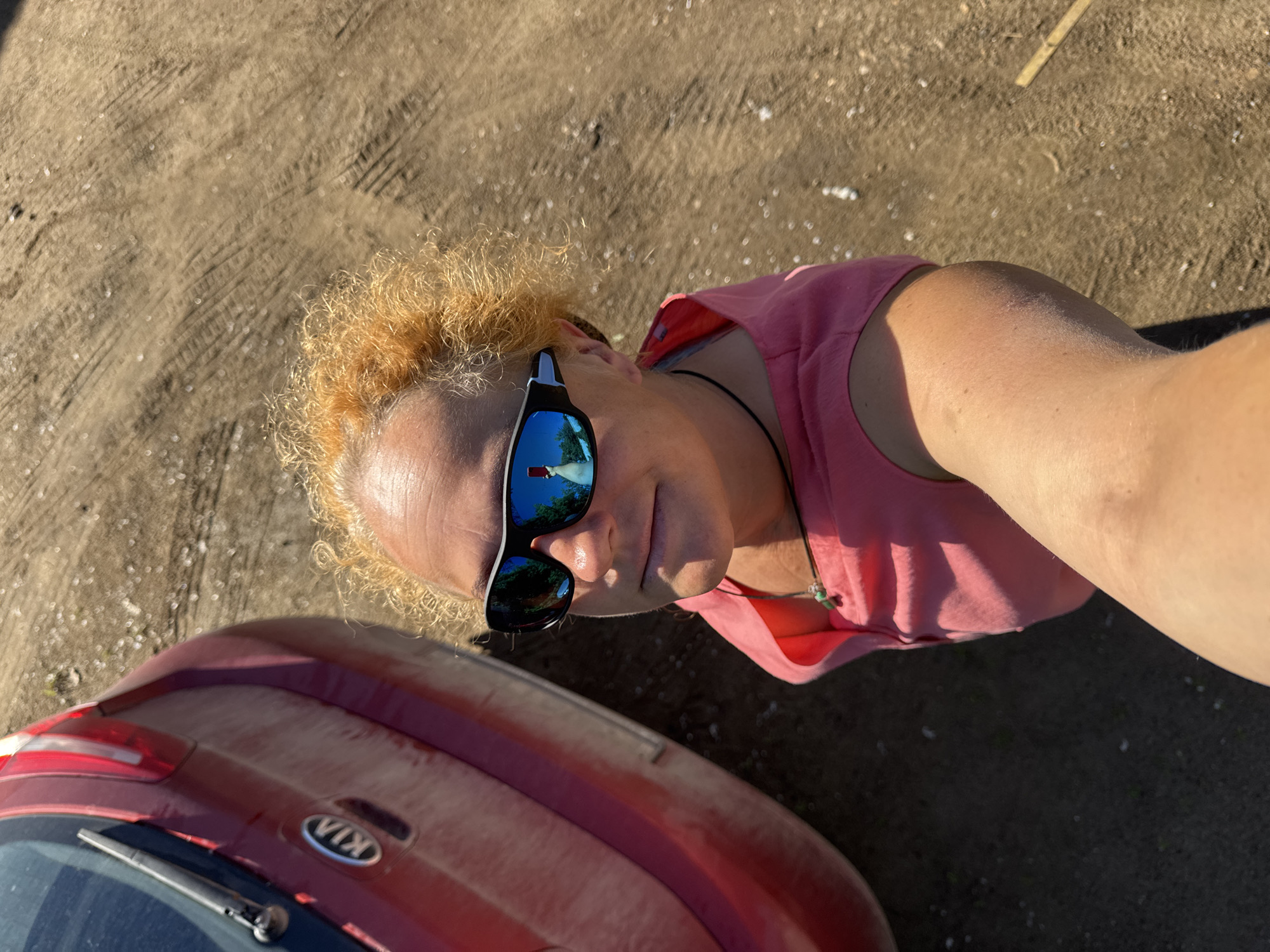 A close-up, overhead selfie shows a person with curly, ginger hair and sunglasses, standing next to the rear of a red Kia vehicle. The person is wearing a pink sleeveless top and a necklace with a green pendant. The sunglasses reflect a person holding a phone, presumably taking the selfie, along with trees and a sky. The ground is dirt with tire tracks and scattered white debris. The scene appears to be outdoors, possibly in a rural or undeveloped area, with no specific landmarks to identify the location within Chaikovsky, Russia. The overall lighting suggests it is daytime.