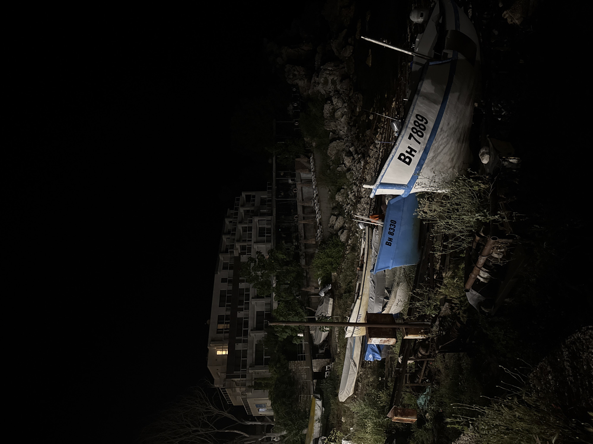 In Tyulenovo, Bulgaria, this image captures a night scene featuring a coastal landscape with a modern building and several boats. The building, likely a hotel or apartment complex, is multi-story with balconies and windows, one of which is lit. The foreground is occupied by a collection of boats resting on what appears to be a rocky or grassy embankment. Some boats are partially covered, while others are exposed. There are no people visible in the image. The environment is dark, with the only light sources being the illuminated window in the building and possibly some ambient light reflecting off the water or boats, creating a subdued atmosphere. The scene suggests a quiet, possibly off-season, coastal setting.
