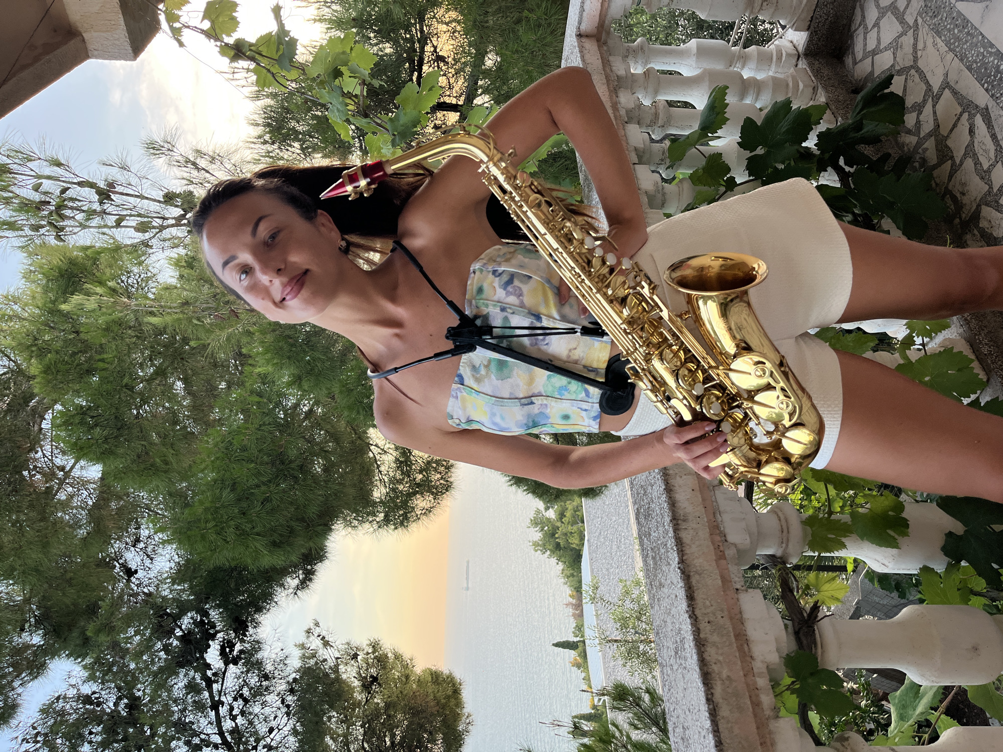 A woman stands on a balcony in Susanj, Montenegro, holding a saxophone. She is wearing a floral print strapless top and white shorts. The balcony has white balusters and a stone railing, with grapevines growing on it. Behind her, the Adriatic Sea is visible with a cargo ship in the distance. Lush green trees frame the scene. The sky is a soft blend of yellow and blue, indicating either sunrise or sunset. The woman is looking at the camera with a slight smile.
