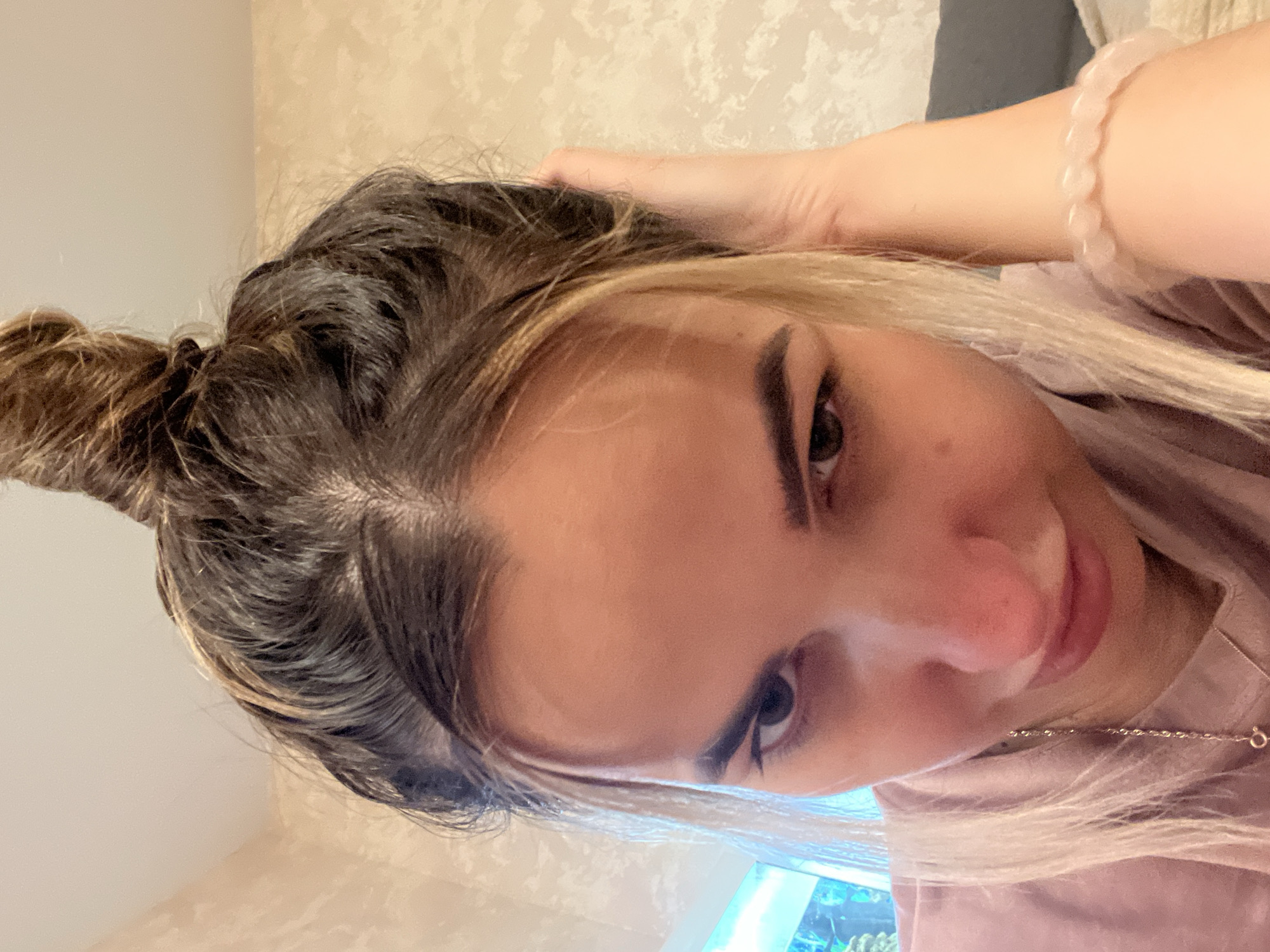 The image is a close-up, slightly downward-facing selfie of a young woman indoors. She has dark brown hair pulled up into a messy bun with some blonde streaks visible around her face. She is wearing a pink shirt and a delicate silver necklace. A pink beaded bracelet is visible on her right wrist. Her left hand is touching her head. The background is a textured, light-colored wall and a glimpse of what appears to be a window with blue light. The overall lighting is warm, and the focus is on the woman's face.