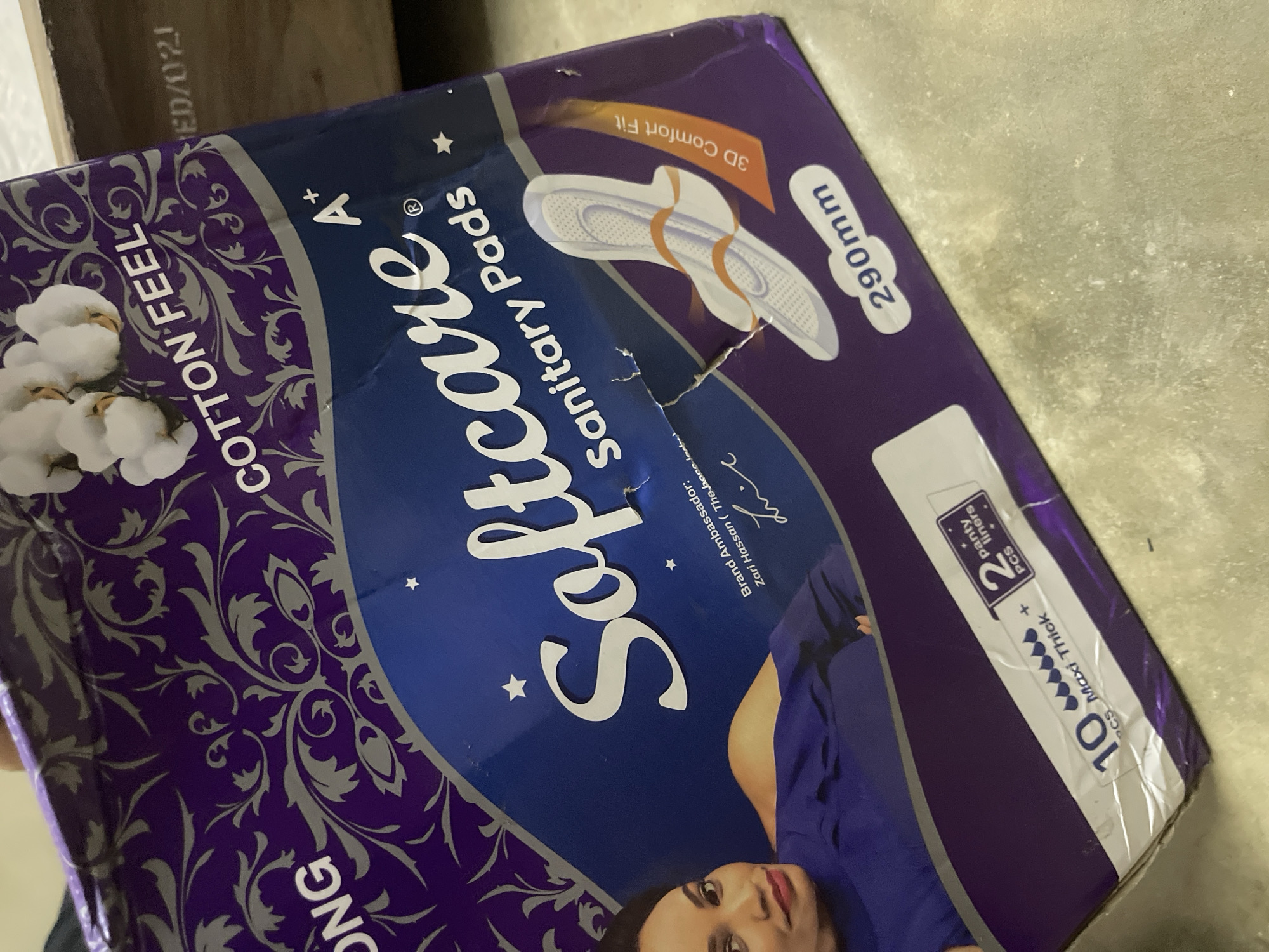 A partially damaged package of "Softcare" brand sanitary pads is angled on a gray surface. The box is primarily blue and purple, featuring cotton flowers and ornamental swirls. Text on the package indicates "Cotton Feel," "Sanitary Pads," "Maxi Thick" with 10 pads, and includes 2 panty liners. The package also indicates "290mm" and "3D Comfort Fit." A person, identified as "Brand Ambassador: Zari Hassan," is pictured on the packaging. The location is implied to be Dar es Salaam, Tanzania, based on information likely associated with the brand.